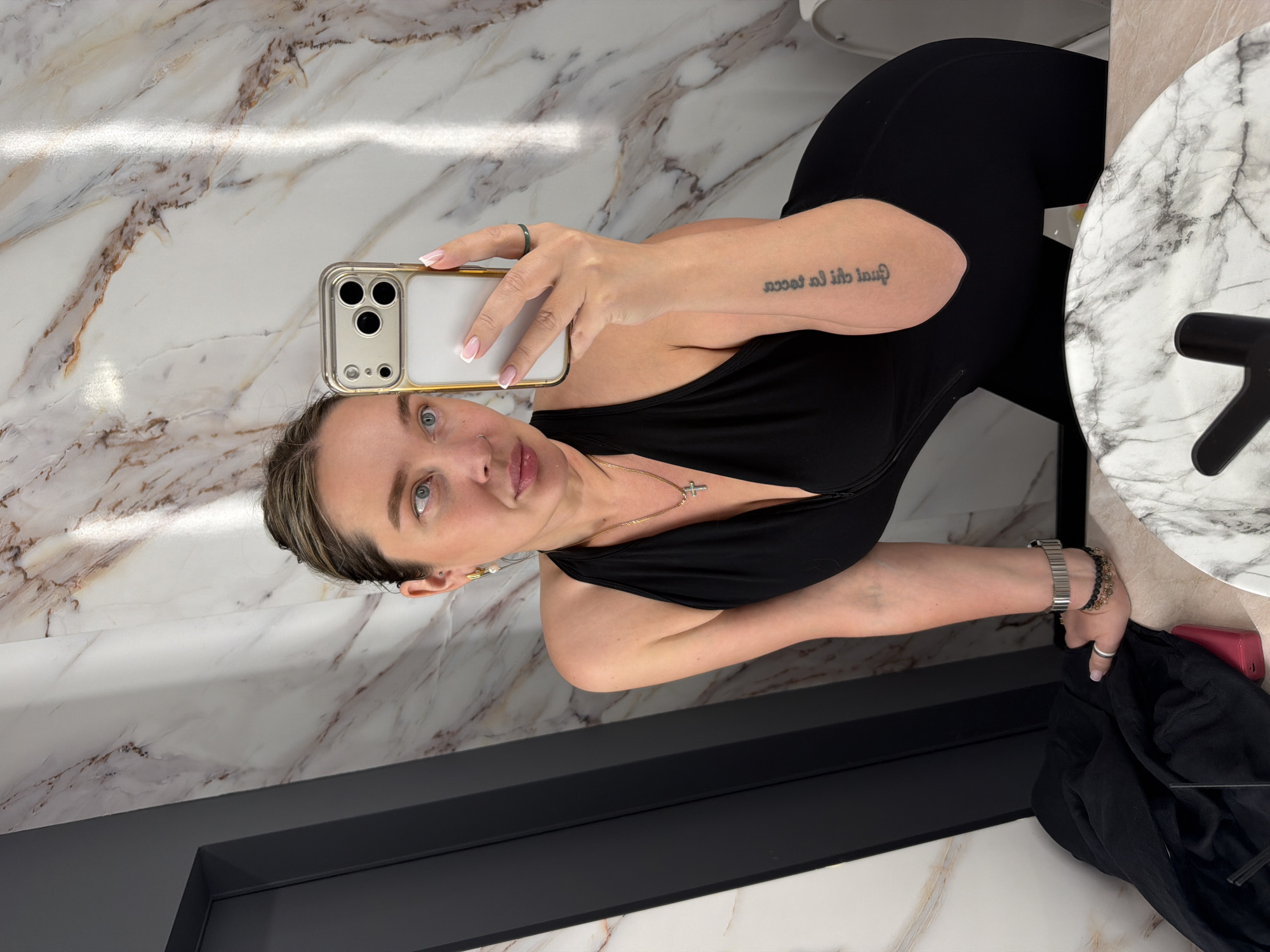 A woman takes a selfie in a bathroom with marble walls and floor. She is wearing a black top and leggings. She is holding a gold iPhone. She has a tattoo on her left arm. She is wearing a necklace with a cross pendant. There is a sink with a marble countertop in the background. A black door is on the left. The location in the city of Moscow, Russia cannot be definitively determined.