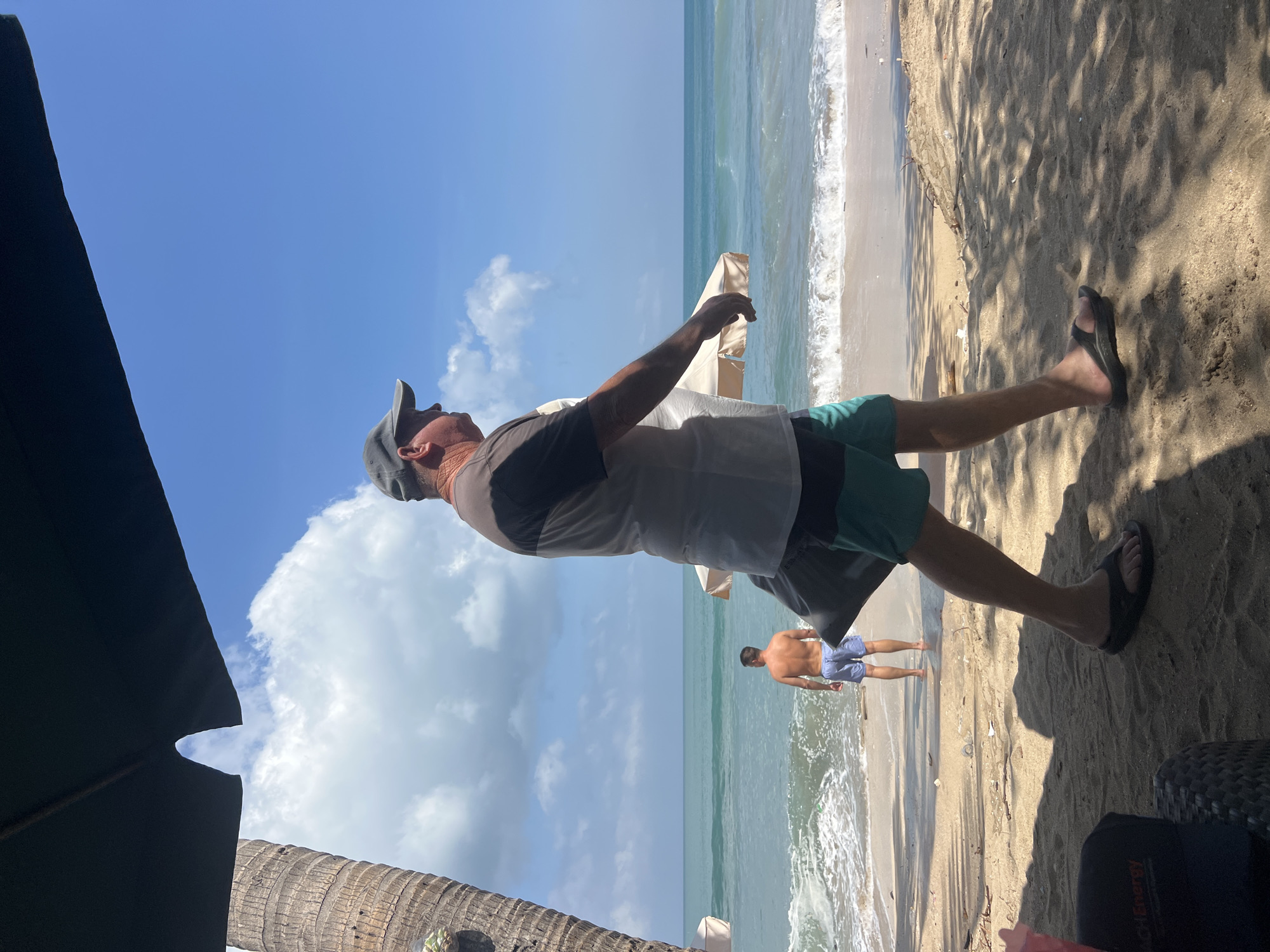A man wearing a cap, a t-shirt with a dark and light gray design, and teal-and-black swim trunks walks along a sandy beach. He carries a dark gray bag in his left hand. The man is facing to the right of the frame. In the background, another man wearing light blue swim trunks stands in the shallow water, facing away from the camera. The ocean has gentle waves breaking on the shore. There are two light-colored beach umbrellas and a palm tree trunk visible. The sky is blue with scattered white clouds. The foreground is shaded by what appears to be a large umbrella or canopy. Part of a black chair and a black cap with the logo "BEACH ENERGY" are visible in the lower left corner. The scene is consistent with Kuta, Indonesia, a popular tourist beach destination known for its wide sandy shores and surf.