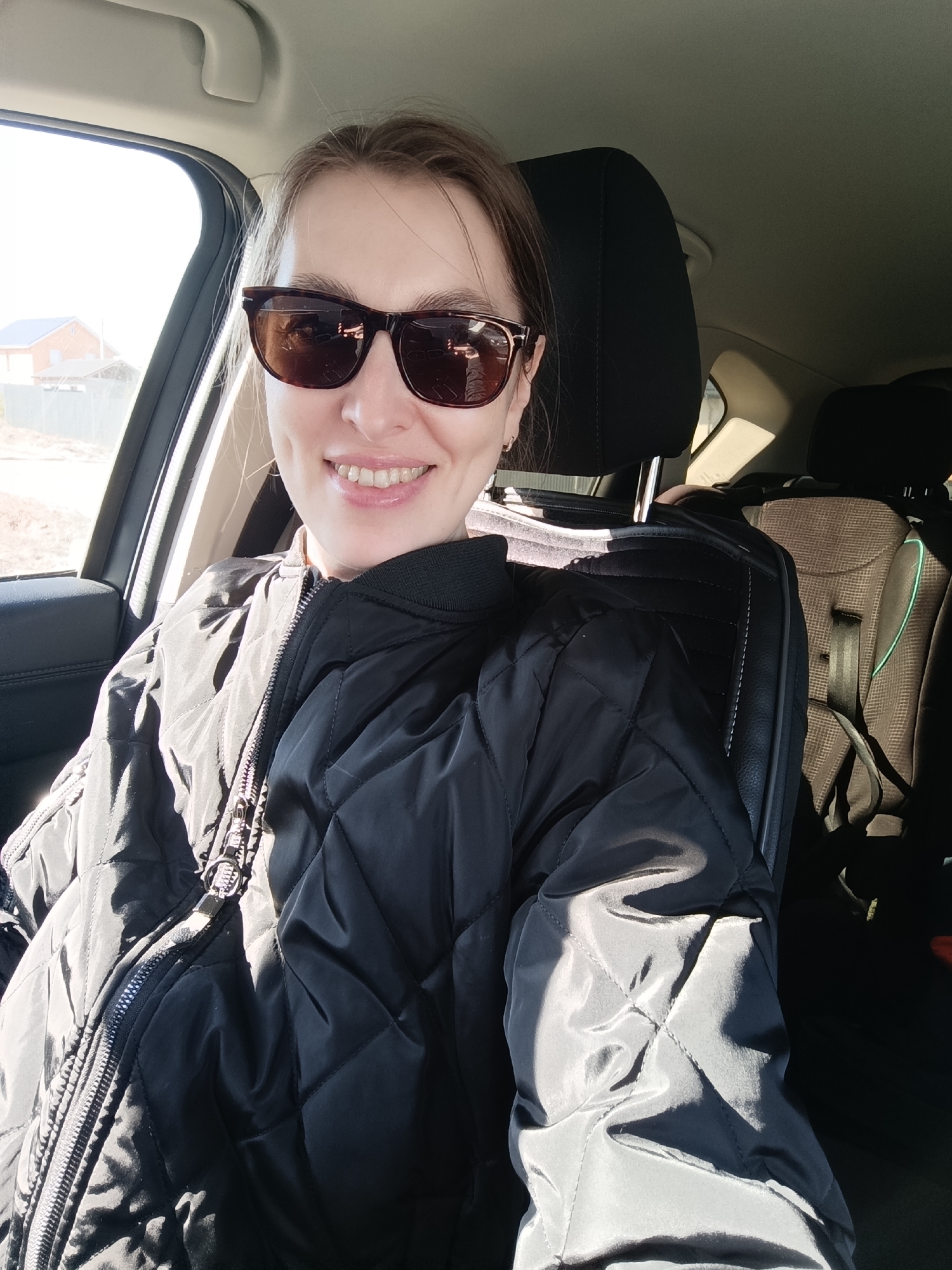 The image is a selfie taken by a woman inside a car, likely in Chaikovsky, Russia, based on the visible building in the background. She is wearing sunglasses and a black quilted jacket. She is smiling. A child safety seat is visible in the back of the car. Outside the car window, there is a glimpse of a house or building. The scene appears to be daytime, with bright sunlight.