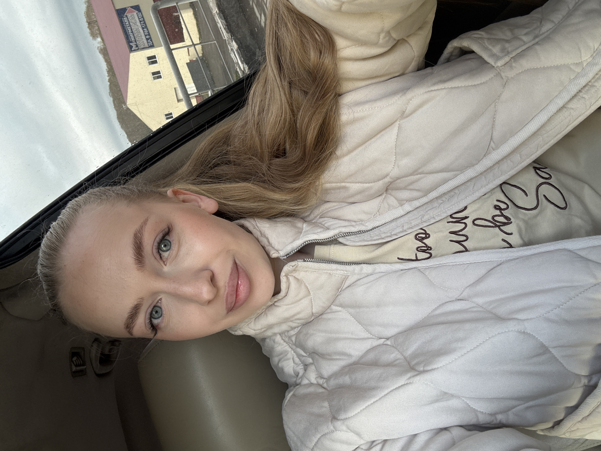A young woman is taking a selfie. She is in a car, likely in Petropavlovsk-Kamchatsky, Russia, based on the Russian signage visible through the car window on a building. The weather is overcast. She is wearing a light-colored, quilted jacket over a light-colored top. She has blonde hair and is looking at the camera with a slight smile. The interior of the car is light-colored.