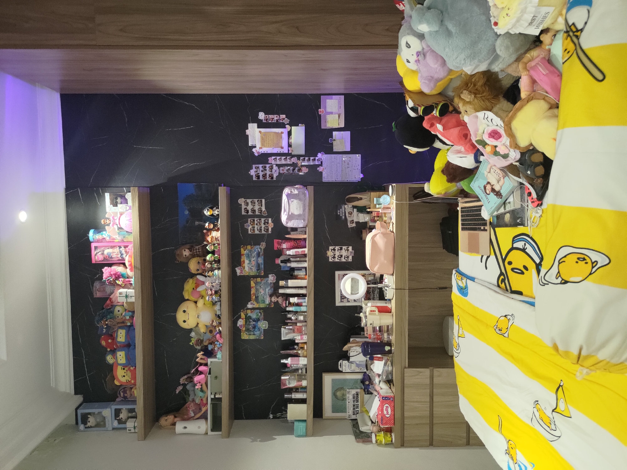 The image shows a bedroom with shelves filled with dolls, plush toys, and cosmetic products. There is a desk with various items on it, including a laptop and a makeup mirror. A bed with Gudetama-themed bedding is visible in the foreground. The room is decorated with photos and other personal items. The scene is likely in Depok, Indonesia.