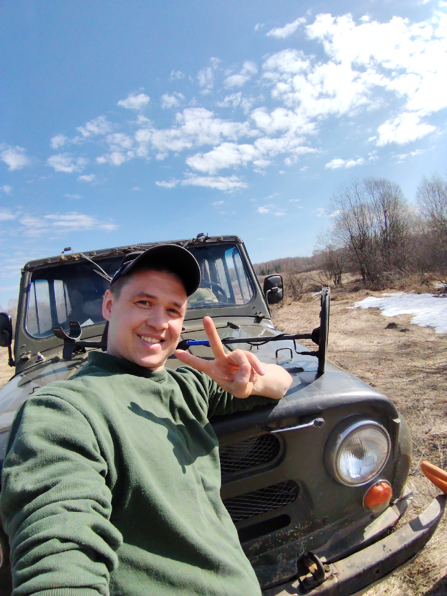 A young man, likely in his 20s or early 30s, is captured in a bright outdoor selfie or photograph. He wears a dark green long-sleeved fleece sweater and a dark baseball cap. The man is smiling widely, showing his teeth, and holds up his right hand in a peace sign gesture. His left arm rests on the hood of a rugged, dark green off-road vehicle, possibly a UAZ-469, which fills a significant portion of the lower frame. Visible parts of the vehicle include its front hood, windshield, driver's side mirror, and a circular headlight with an orange turn signal.
The setting is an open, undeveloped landscape in what appears to be Markovskii, Russia. The ground is covered with dry, light brown grass and dirt. Patches of snow are visible on the ground to the right of the vehicle, suggesting either late winter or early spring. Sparse, bare deciduous trees without leaves line the horizon in the background. Above, the sky is a vibrant blue with scattered white, fluffy clouds, indicating clear and sunny weather. The strong sunlight suggests it is daytime, likely late morning or early afternoon. The man's cheerful expression conveys a sense of happiness or enjoyment of the outdoor environment. There is no discernible text visible in the image.