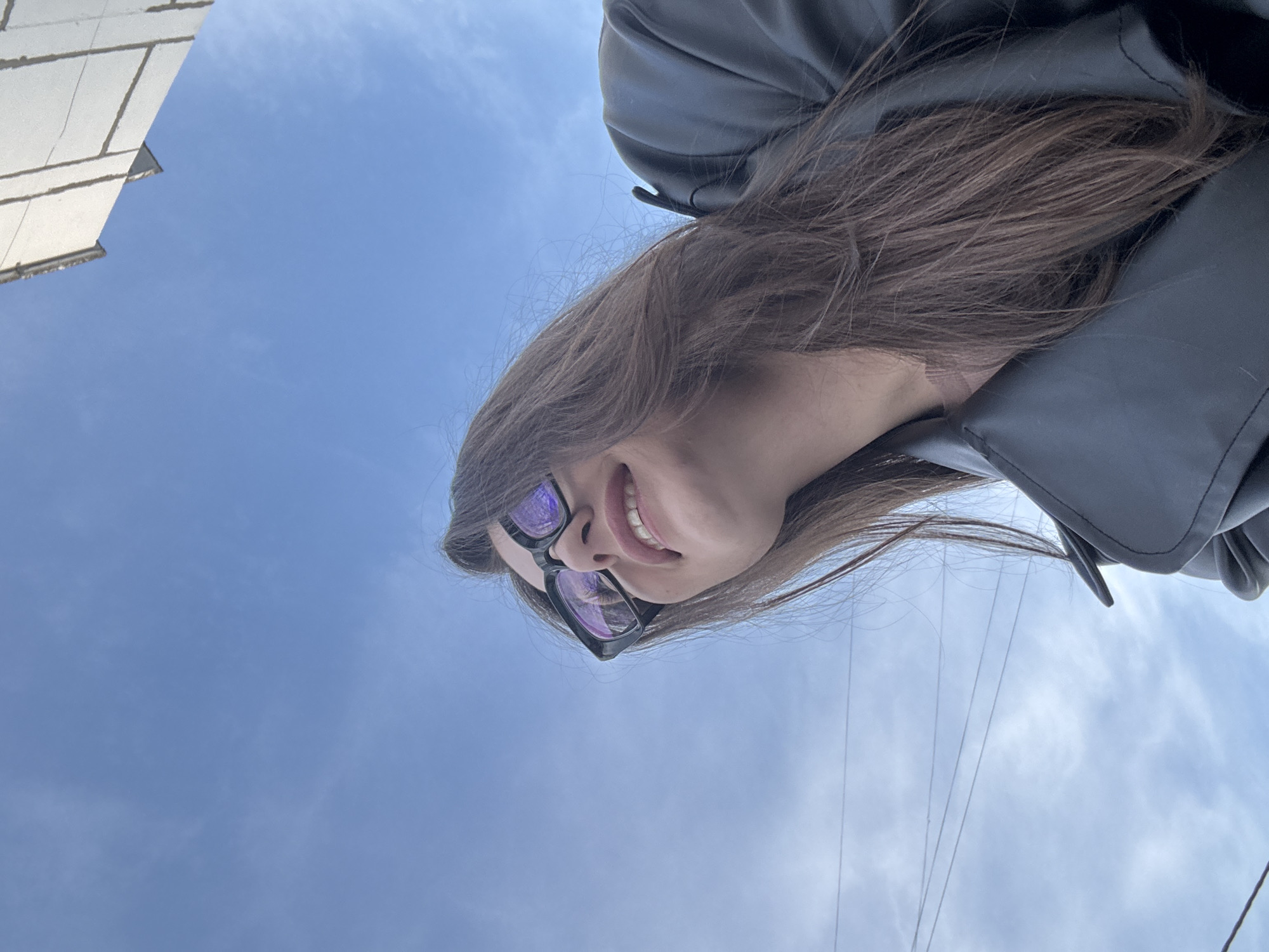 The image displays a female individual situated outdoors in Chaikovsky, Russia, captured from a low-angle perspective. She is looking upwards and slightly to her left, with her mouth open in a smile, revealing her upper teeth. She has long, dark brown hair and is wearing black-framed sunglasses, which show purple reflections in their lenses. Her attire includes a dark-colored (likely black or dark grey) jacket or coat, visible on her upper torso and shoulders. The background primarily consists of a clear, light blue sky featuring scattered wispy white clouds. In the upper right quadrant, a portion of a light-colored building with visible panel lines is present. Multiple thin, dark horizontal lines, consistent with utility wires, traverse the sky in the mid-left and lower-left areas of the frame.