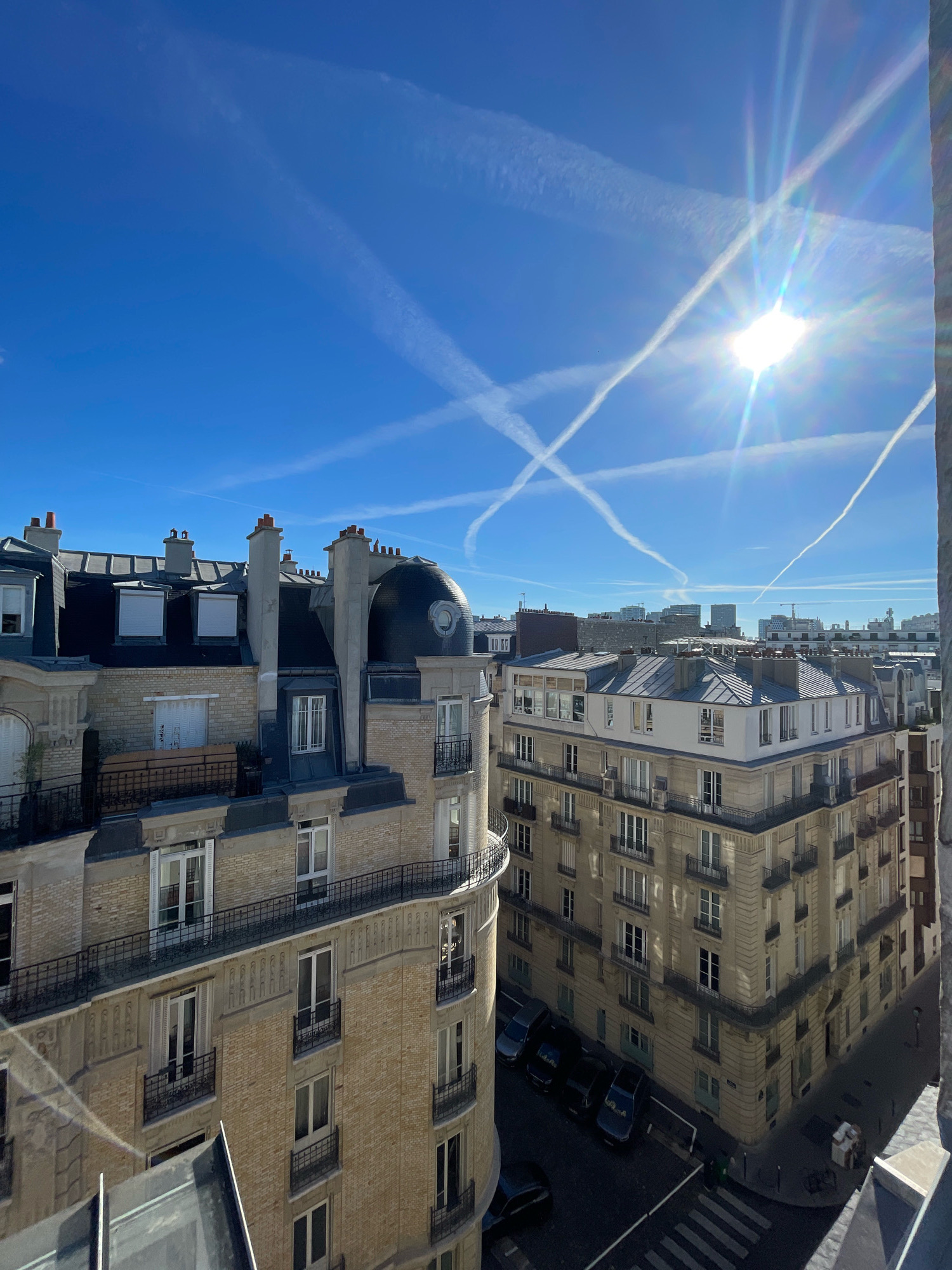 An elevated, eye-level, medium shot captures a clear blue sky filled with multiple contrails from airplanes, forming an 'X' pattern intersecting near the sun, which is a bright orb with lens flare extending outwards. The sun is positioned in the upper right quadrant of the image. Below the sky are the rooftops and facades of Parisian buildings.
In the foreground, to the left, is the upper portion of a pale yellow brick building with a dark gray mansard roof, adorned with dormer windows and chimneys. A decorative balcony with wrought-iron railings runs along one section of this building.
To the right of this building is another, larger building, also made of pale yellow brick, with a gray mansard roof. This building has numerous windows with balconies, also featuring wrought-iron railings. Its facade is decorated with architectural elements. The sunlight casts strong shadows on this building.
In the lower right corner, a street is visible, with a crosswalk and several parked cars. The street is lined with buildings, and the shadows suggest it is daytime. The overall scene depicts a typical urban landscape in Paris, France, under a bright, clear sky.