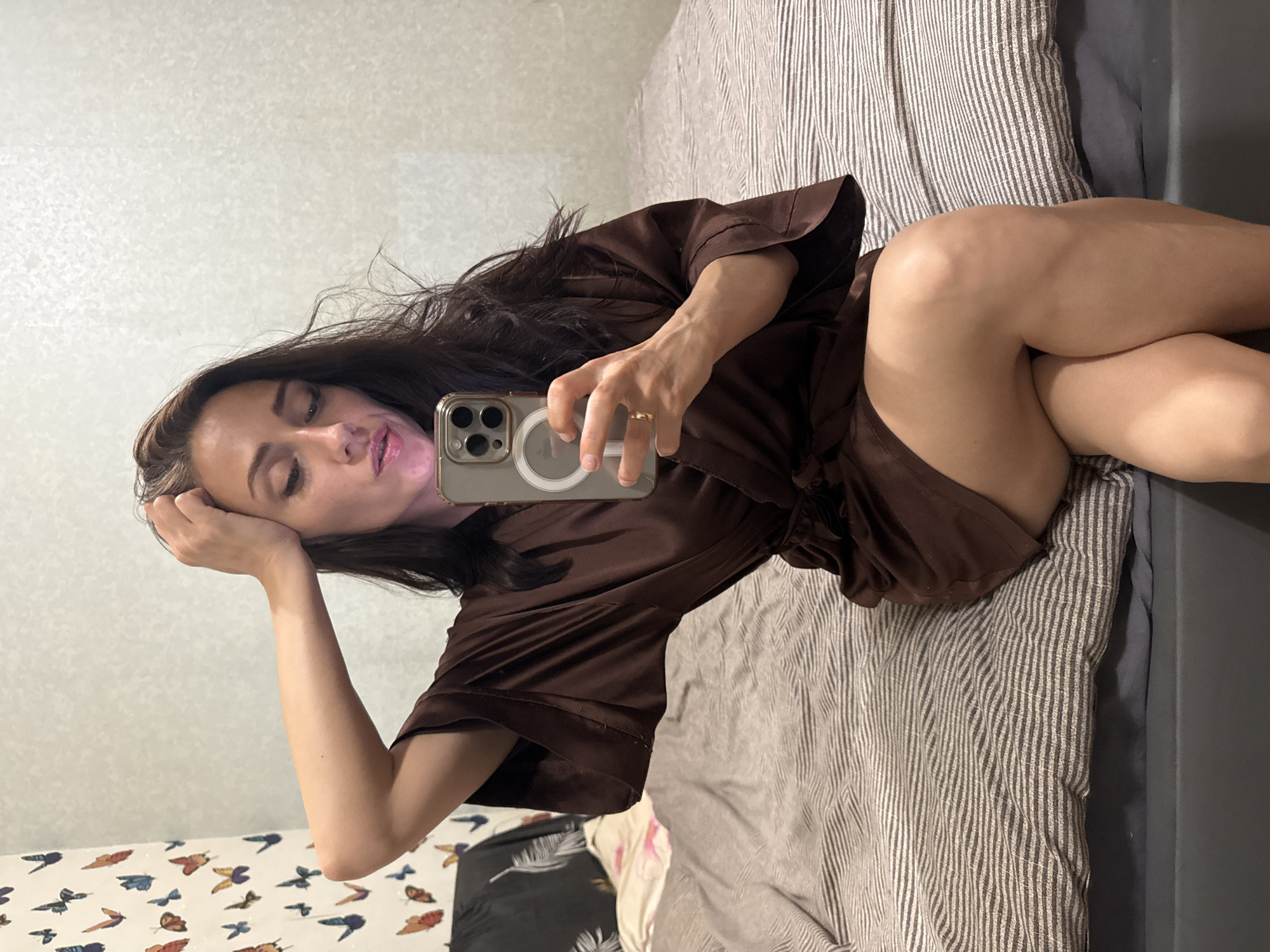 The image shows a woman in a brown satin robe lying on a bed. She is holding a smartphone, taking a selfie. Her left arm is resting on her head, and her right arm is bent at the elbow, holding the phone. Her eyes are closed, and her lips are slightly parted, giving the impression of relaxation or a playful pose. The bed has striped gray and white bedding. In the background, the wall has a subtle textured pattern, and part of a decorative wallpaper with butterflies is visible near the bottom of the frame. The lighting suggests it might be daytime, possibly in the morning or afternoon, given the natural light reflecting on the phone screen. The overall mood is intimate and casual, capturing a personal moment. No text is visible in the image. The location context provided is Saint Petersburg, Russia.