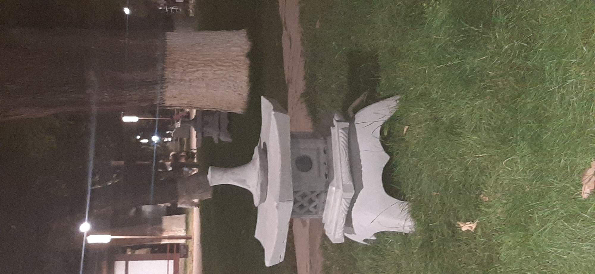 The image shows a nighttime scene with an outdoor setting. The main subjects are several light-colored, sculptural objects lying on a patch of green grass. These objects appear to be made of a solid material like concrete or stone and have a geometric, somewhat abstract design. One of the objects resembles a stylized wing or fin. In the background, there is a body of water with reflections of lights. There are also streetlights and possibly building lights visible, suggesting an urban or park-like environment. Some of the crops show blurred figures of people in the distance near the water's edge and near some structures. The lighting is dim, indicative of nighttime, and the weather appears to be clear, given the reflections on the water. No text is visible in the image. The overall impression is one of a public space at night, with artistic elements displayed outdoors.