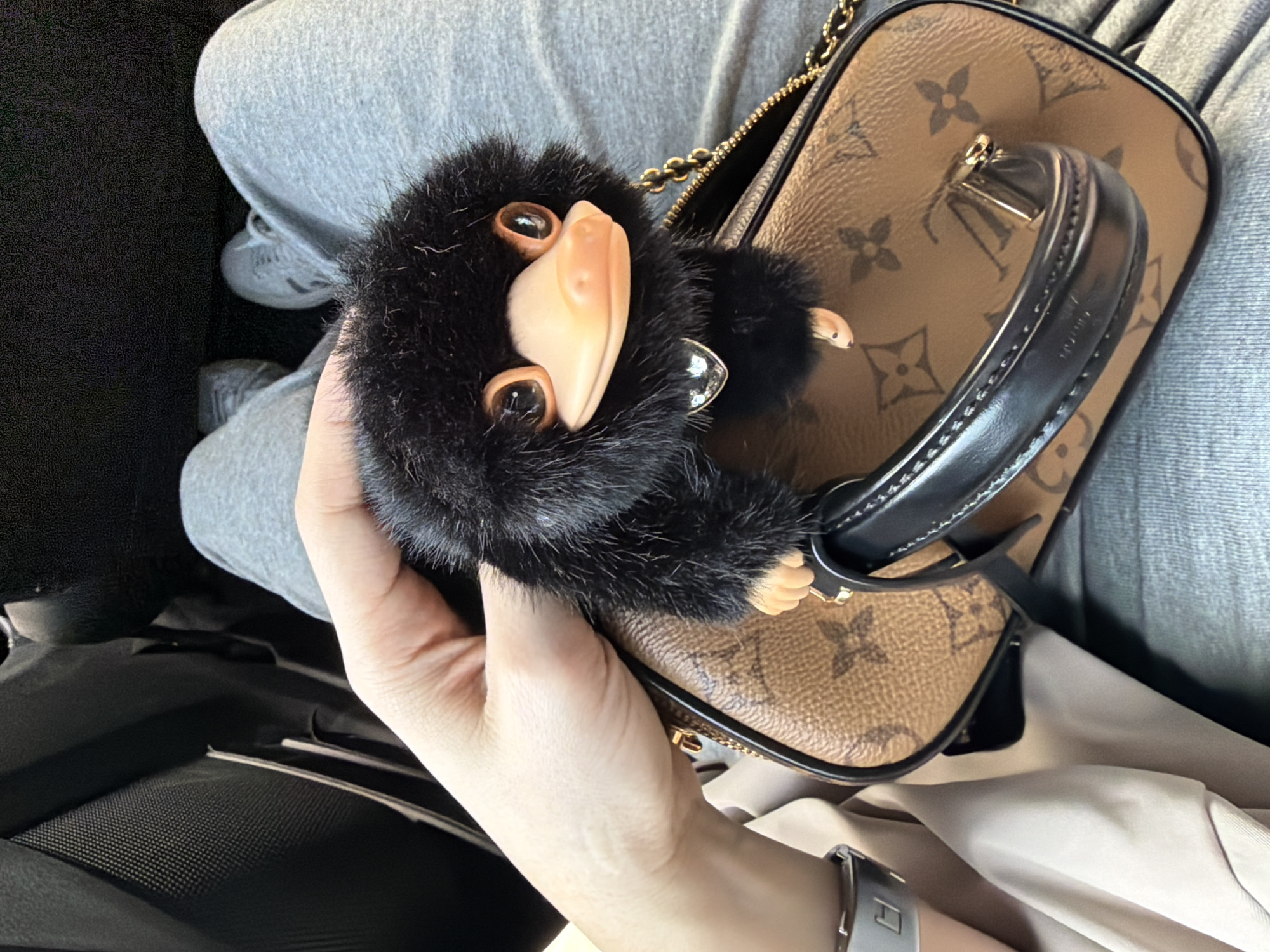 A high-angle close-up shot shows a hand holding a Louis Vuitton handbag with a Niffler charm attached. The handbag is brown with a dark brown monogram pattern and black leather handles and trim, accented with gold hardware. The Niffler charm is black and furry with a pink beak. The hand holding the bag has a silver watch on the wrist. The background consists of the person's legs wearing grey sweatpants and a glimpse of a grey sneaker. Parts of a car interior are visible as well. The location is the Chaoyang District, China.
