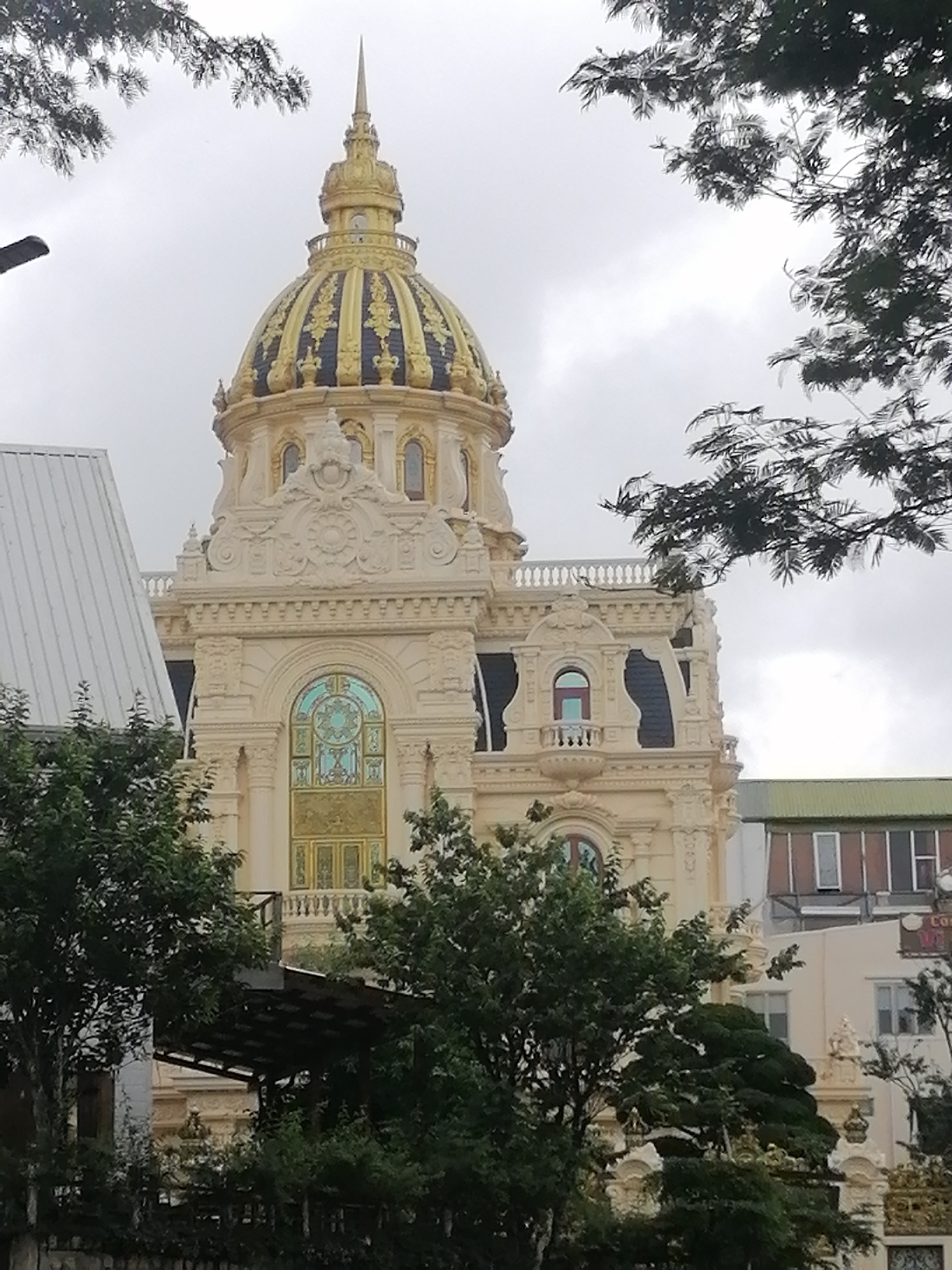 The image captures a grand, ornate building featuring a prominent golden dome with a slender spire, set against an overcast sky. The central structure is cream or light beige, adorned with intricate architectural details, carvings, and moldings, characteristic of European classical or Baroque styles, common in French colonial architecture. The dome itself is multi-tiered, predominantly gold with dark, possibly blue or black, decorative patterns.
Below the dome, the building's facade showcases arched windows, some with stained-glass-like designs in blue and gold, and a small balcony on the right side. Lush green trees and foliage are scattered in the foreground and frame the top of the image, partially obscuring the building. To the left, a modern structure with a sloped white metal roof stands in contrast to the ornate central building. In the background on the far right, another, plainer building with a green roof and visible windows can be seen, with a sign containing partially obscured, unreadable text.
The scene depicts a static architectural view under cloudy, overcast conditions, suggesting daytime without direct sunlight. No specific activity or people are visible. The elaborate design of the main building, particularly its dome and decorative elements, points to a significant landmark, fitting the architectural landscape of a location like Da Lat, Vietnam.