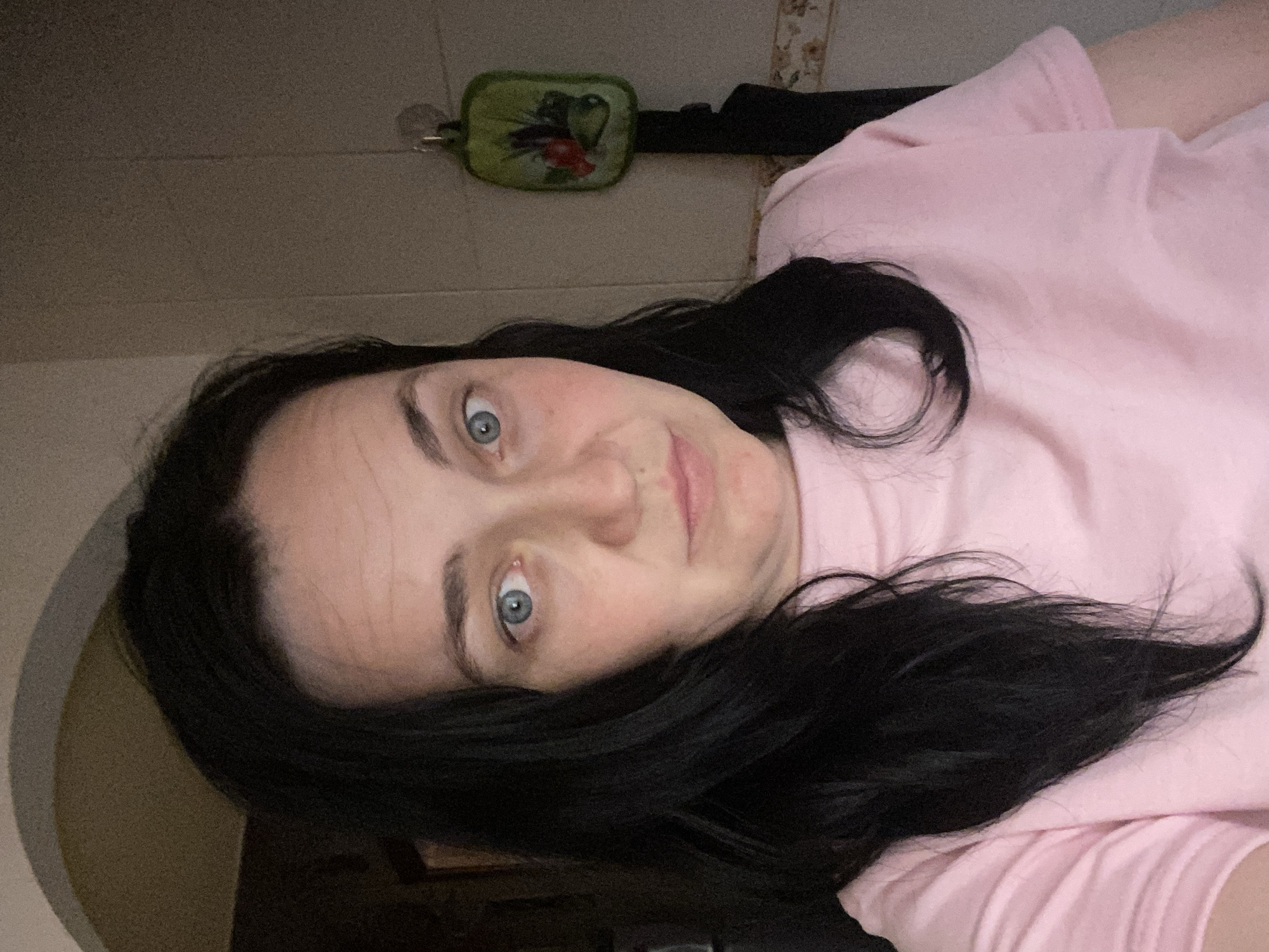 The image is a close-up, selfie-style photograph of a woman with long, dark hair and blue eyes. She is wearing a pink t-shirt. Her facial expression is neutral, with a slight pout. The background is a wall with some kitchenware hanging on it, and an archway. The lighting is dim, creating shadows on her face. The image appears to be taken indoors, likely in a home environment, but no specific city or location details are discernible from the visual information.