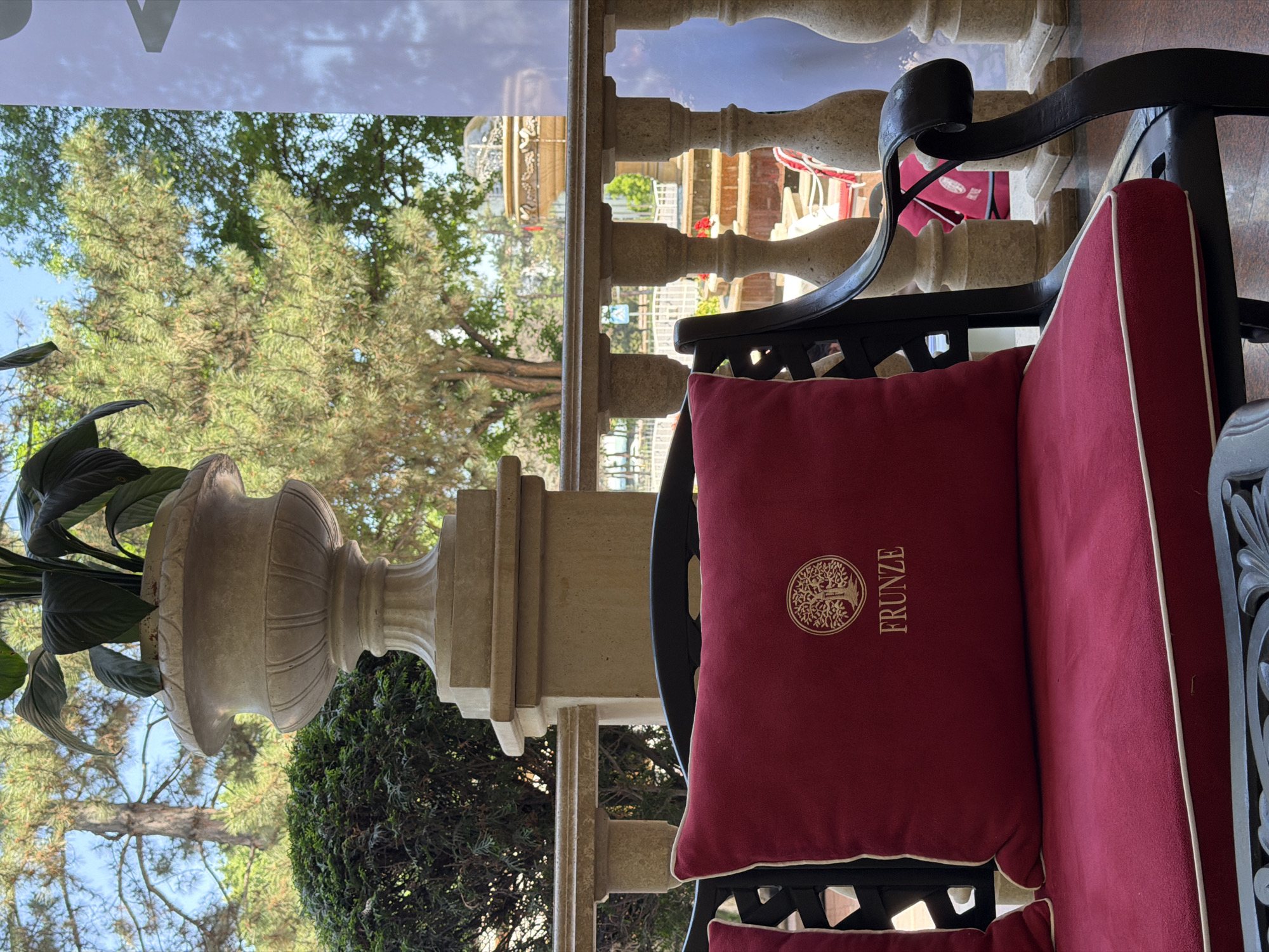 The image captures a scene on a terrace or balcony, likely in a hotel or restaurant, in what appears to be Bishkek City, Kyrgyzstan, given the "FRUNZE" embroidered on a red cushion. The cushion is placed on a black metal chair with red padding along the edges. Behind the chair, there's a stone balustrade, and beyond that, trees and possibly other outdoor seating areas are visible. To the left of the chair is a large, ornate stone flower pot containing plants with large green leaves. The overall setting suggests an upscale outdoor space, designed for relaxation or dining.