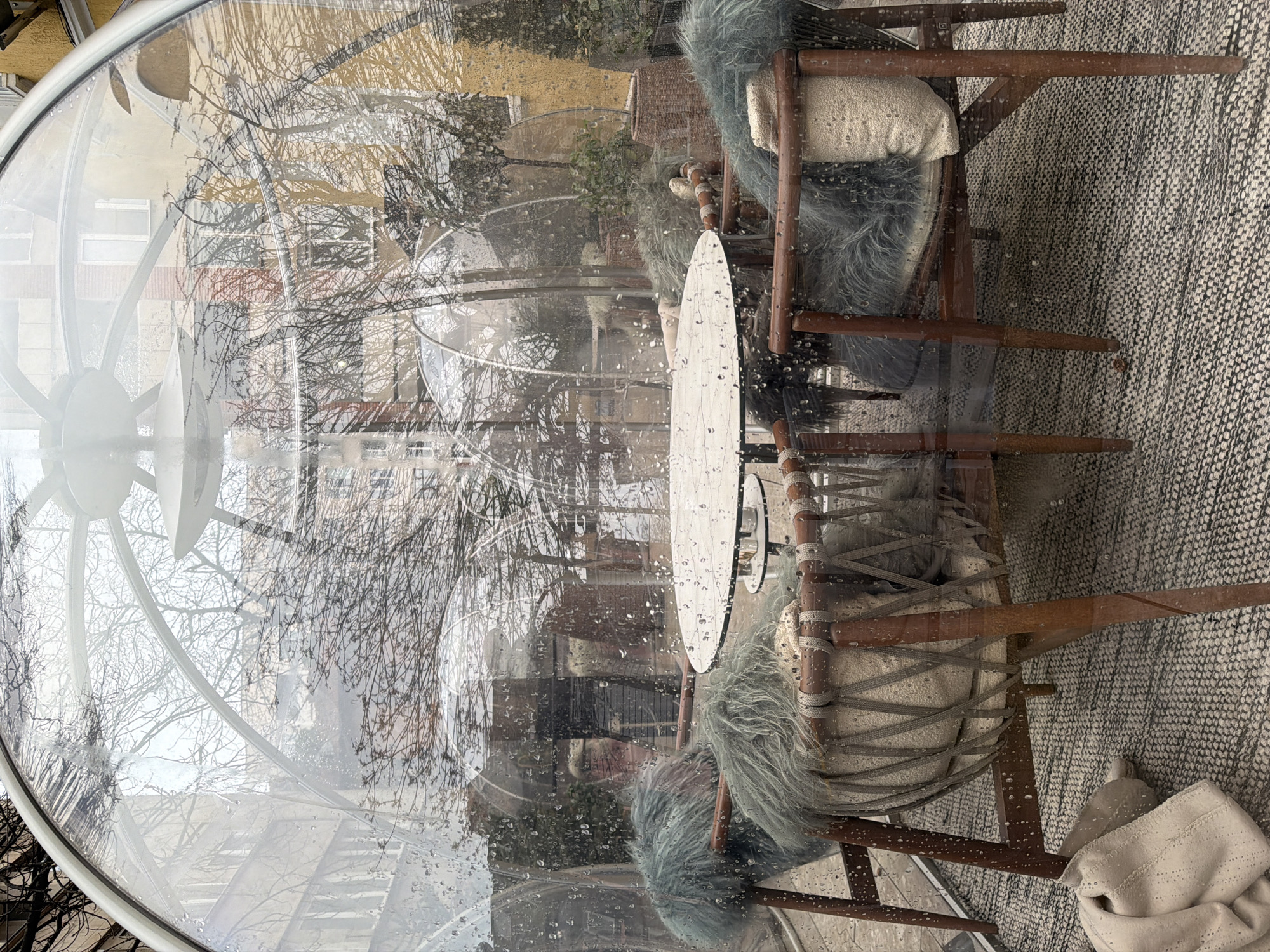 An outdoor urban dining or lounge setting in Sofia, Bulgaria, is depicted from inside a transparent, geodesic-style dome enclosure. Raindrops are visible across the exterior surface of the clear dome.
Centered within the dome is a circular white table with a metallic base. Surrounding the table are four wooden chairs featuring woven backrests and seat bases. Each chair is equipped with a shaggy teal/grey faux fur throw and a cream-colored, textured cushion. A light-colored, folded blanket lies on the patterned black and white woven rug covering the floor inside the dome.
No individuals are present within the enclosure.
Through the transparent dome and its reflections, an urban environment is visible. This includes multiple multi-story buildings of varying architectural styles and colors, such as cream, beige, yellow, and red brick. Numerous bare deciduous trees, indicative of a dormant season, are scattered throughout the background. Reflections of at least one other similar transparent dome structure are also discernible in the glass. The ambient lighting suggests an overcast sky.