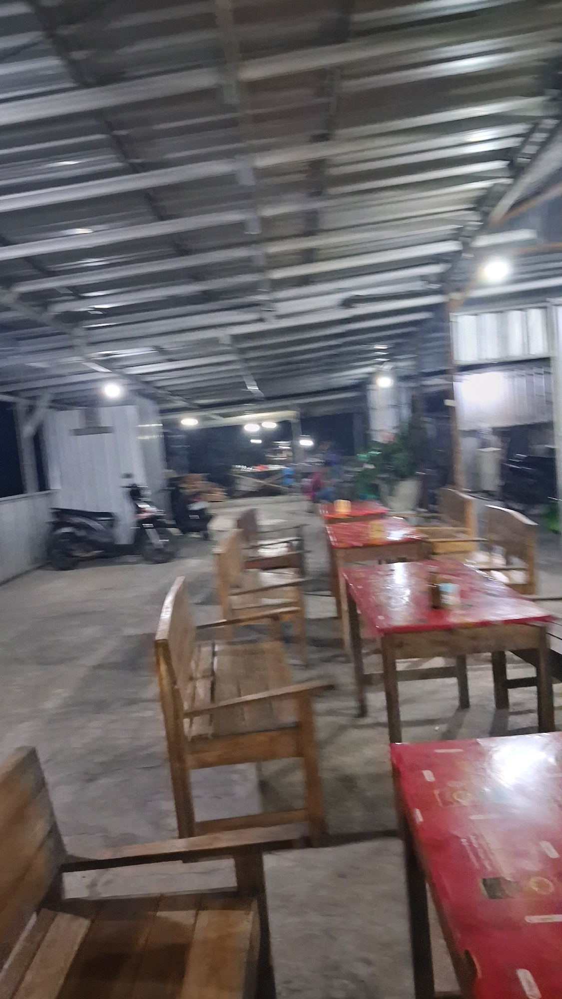 The image depicts an outdoor seating area, likely a food stall or restaurant, under a metal corrugated roof supported by metal beams and poles. The floor is made of concrete. Several wooden tables with red tops and matching wooden chairs are arranged throughout the space. In the background, a motorcycle is parked next to a white structure. Further in the distance, more tables and chairs are visible, along with some vegetation and distant lights. The scene is dimly lit, suggesting it might be evening or early morning. The location is identified as Berbah, Indonesia.