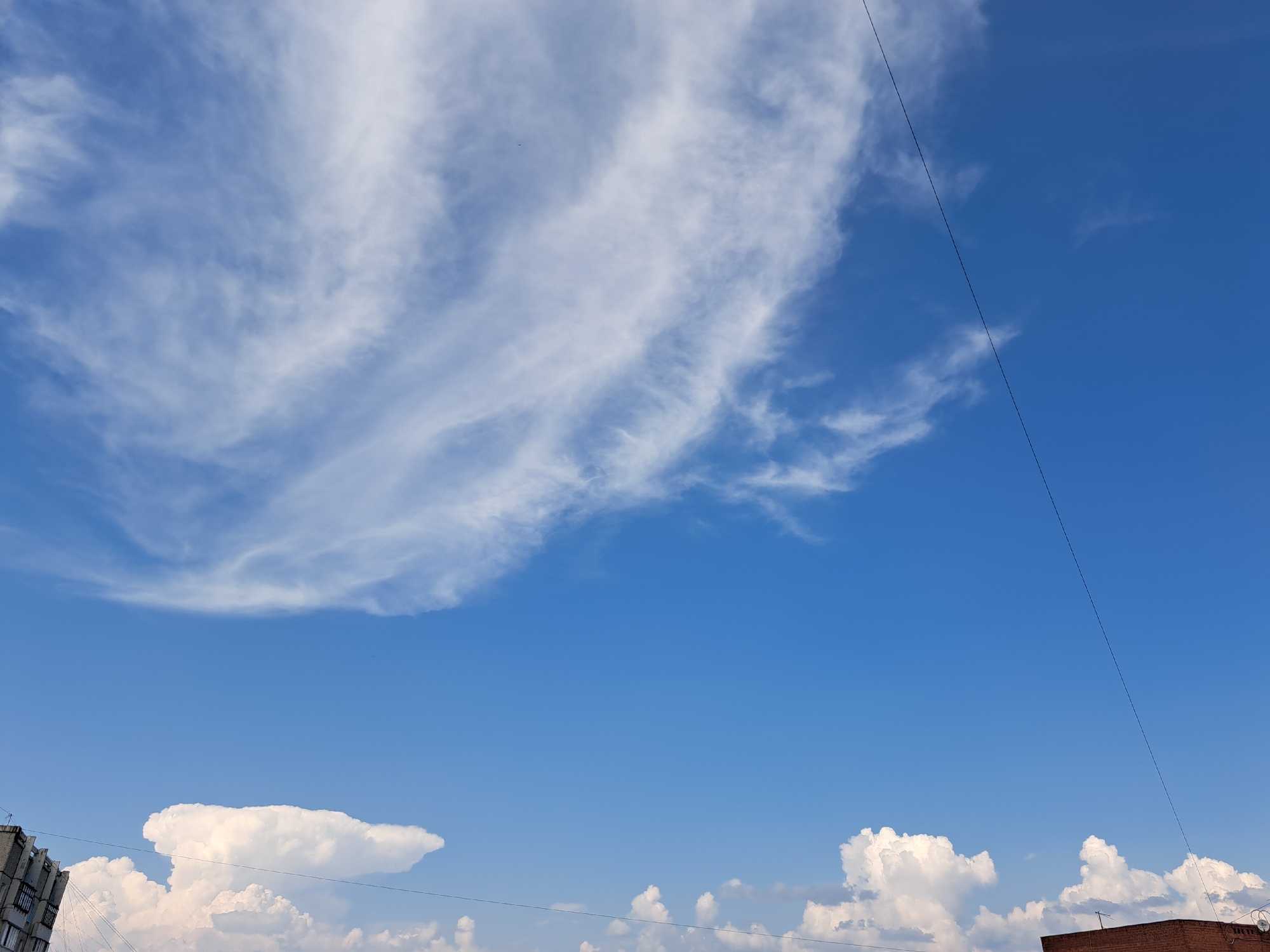 The image depicts a bright blue sky with scattered white clouds. In the lower portion of the image, the rooftops of buildings are visible. One building on the left appears to be a multi-story residential complex, while another on the right is a brick building with a visible antenna. A thin black line, likely a power line, stretches diagonally across the sky. The scene is characteristic of an urban environment on a clear day. The image appears to be taken in the city of Irkutsk, Russia.