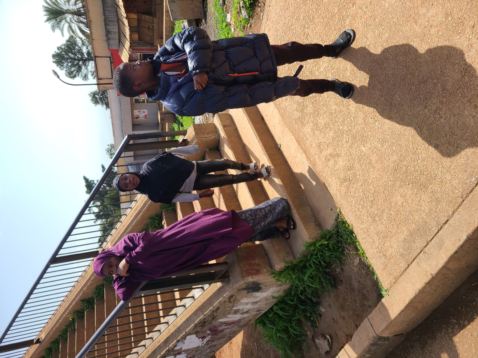 In Gura Top, Nigeria, three young people are positioned on and around a concrete staircase with metal railings. The individual on the left, wearing a purple hijab and patterned pants, leans on the railing, looking down. To their right and slightly down the stairs, another person in a black hijab and grey top is looking forward. The third individual, standing on the ground in front of the stairs and facing right, wears a dark blue puffer jacket and dark pants. The environment suggests an outdoor urban setting with buildings and palm trees visible in the background. The ground is a light brown, gravelly texture, and some green vegetation is present along the base of the stairs and in the background. The scene is illuminated by bright daylight, casting distinct shadows.