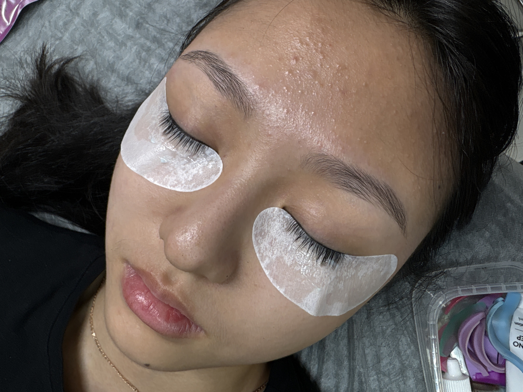 A close-up, top-down shot captures a woman lying on her back, eyes closed, in what appears to be a beauty salon setting. White patches are applied under her eyes, likely for an eyelash extension procedure. Her skin is fair, with visible pores and some small bumps on her forehead. Her eyebrows are well-defined and groomed. She has dark hair. The background includes a grey fabric surface and, to the right, a clear plastic container holding what appears to be tools and materials used for the beauty treatment. A gold chain necklace is visible. The location is Perm, Russia.