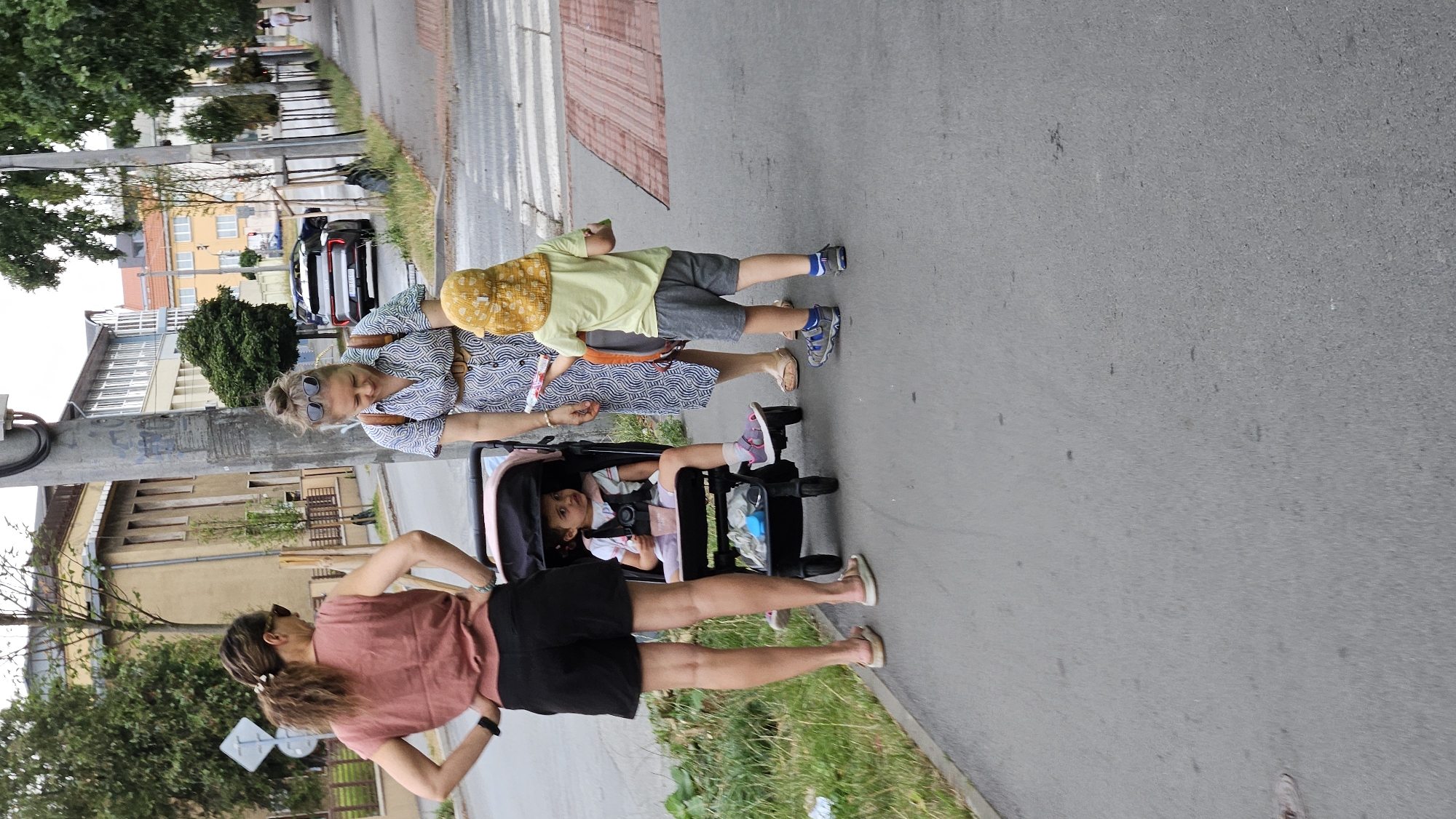 Two adult women and two young children are on a paved sidewalk next to a street. One woman, on the left, wears a pink sleeveless top and black shorts, with her hands on her hips. She is looking down and to her right. Next to her is a stroller with a young child sitting inside, facing forward. The child wears a light pink top and light pink sandals. The second woman, on the right, wears a blue and white patterned dress and is looking down at the child standing in front of her. This child wears a yellow hat, a light yellow t-shirt, and grey shorts. The child is holding a red and white object. Behind the people, a street with a crosswalk is visible, with a parked car and trees lining the street. Buildings can be seen in the background. The scene appears to be in a city.
