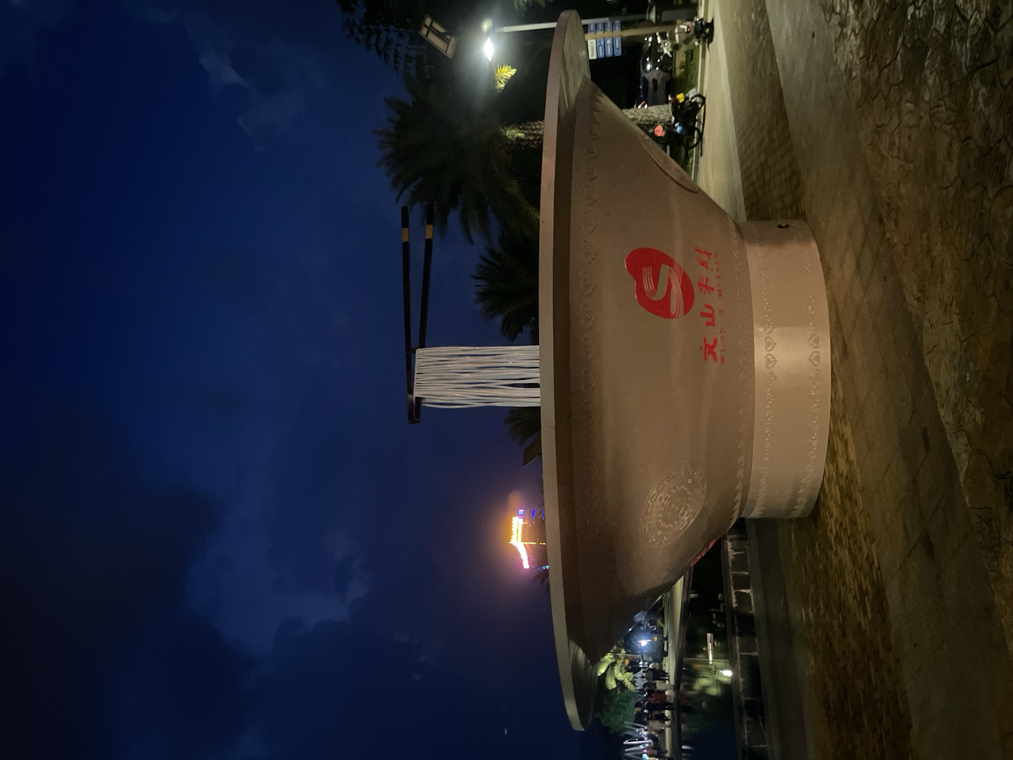 An oversized bowl of noodles, with chopsticks suspended above, is displayed outdoors at dusk or night in a city. The bowl is pale pink with red Chinese characters and a logo on its side, as well as decorative white patterns around its base. The noodles are white and appear to be made of some sort of rope or fabric. Behind the noodle bowl, palm trees are visible against a dark blue sky, with clouds illuminated by distant lights. In the background, a brightly lit building with orange signage stands out. People are gathered near a body of water to the left of the noodle bowl, and vehicles are parked to the right. The ground is paved with interlocking stones.