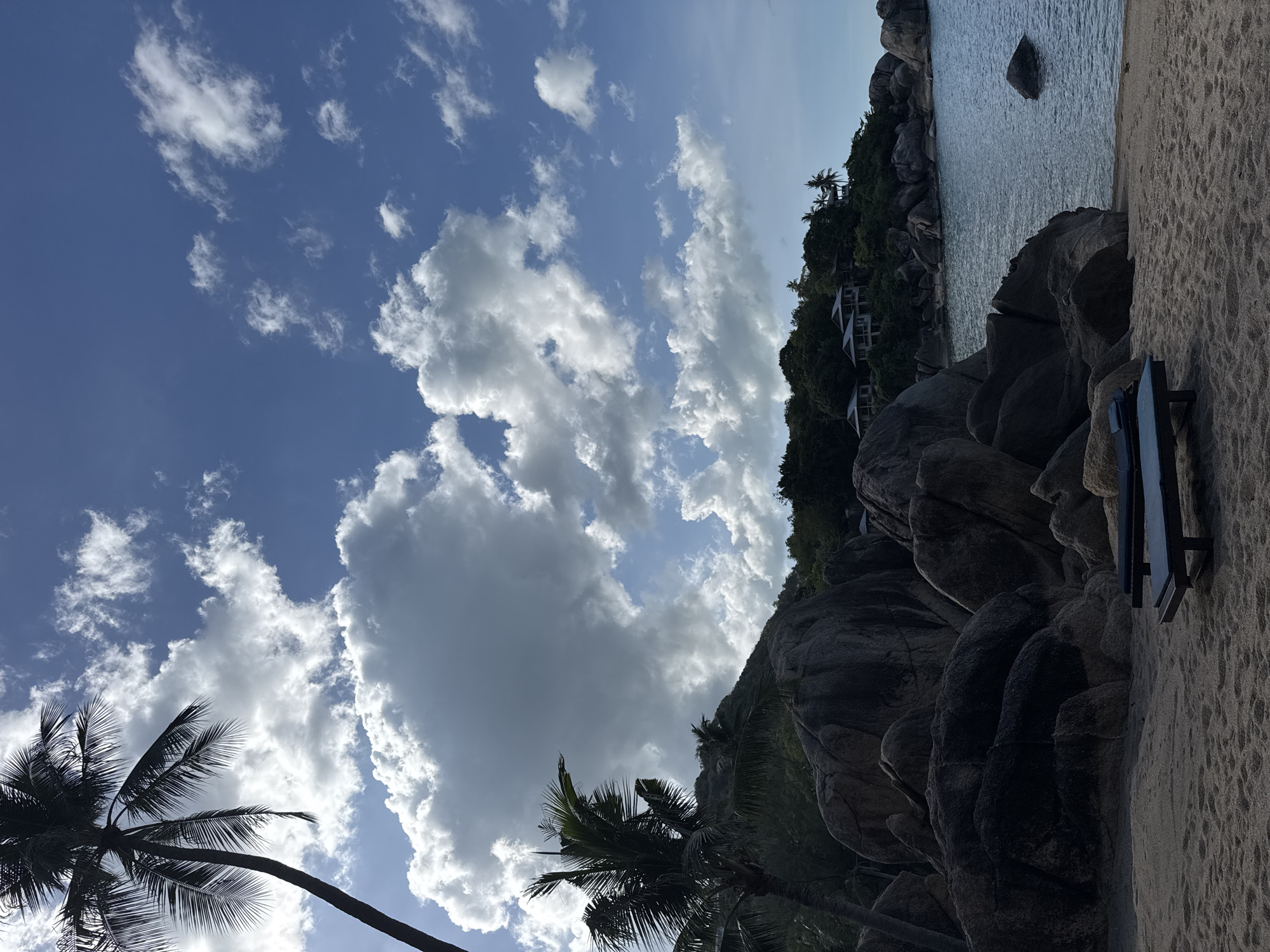 The image shows a coastal scene in Baan Chaweng Noi, Thailand. The view is oriented with the sky above and the beach and ocean to the right. The sky is blue with scattered cumulus clouds, some large and dense, others smaller and more diffuse. A palm tree with drooping fronds is visible in the lower left of the frame. To the right, large granite boulders form a rugged coastline. Vegetation, including trees and shrubs, covers the rocks. White buildings are nestled among the trees on the hillside. The ocean is visible beyond the rocks, with a small rocky island in the distance. A dark object resembling a beach chair or similar furniture is on the sand near the rocks in the right portion of the image. The sand is light in color and appears dry.