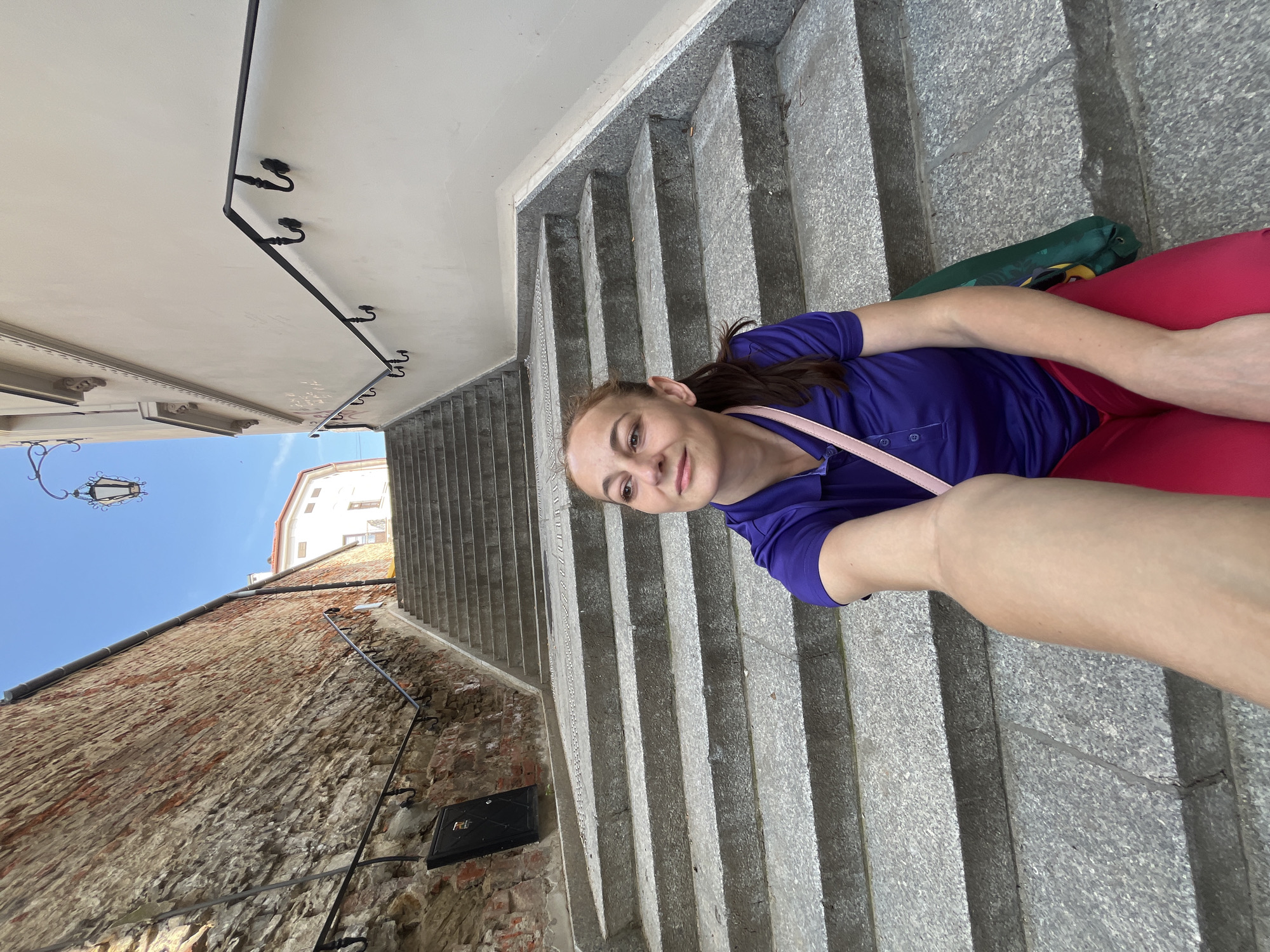 A medium shot shows a woman sitting on stone stairs, looking to her left. She has medium brown hair pulled back in a ponytail, and is wearing a purple polo shirt and bright pink pants. A pink strap from a bag is visible over her shoulder. The stairs are made of light gray stone and appear to be outdoors, leading up between two buildings. To the left is a rough, reddish-brown brick wall with some dark fixtures. To the right is a smooth, light-colored building wall with a black metal handrail. A street lamp hangs from a pole extending from the right building. The sky is a clear, bright blue. The woman's left arm is extended towards the camera, holding it for a selfie.