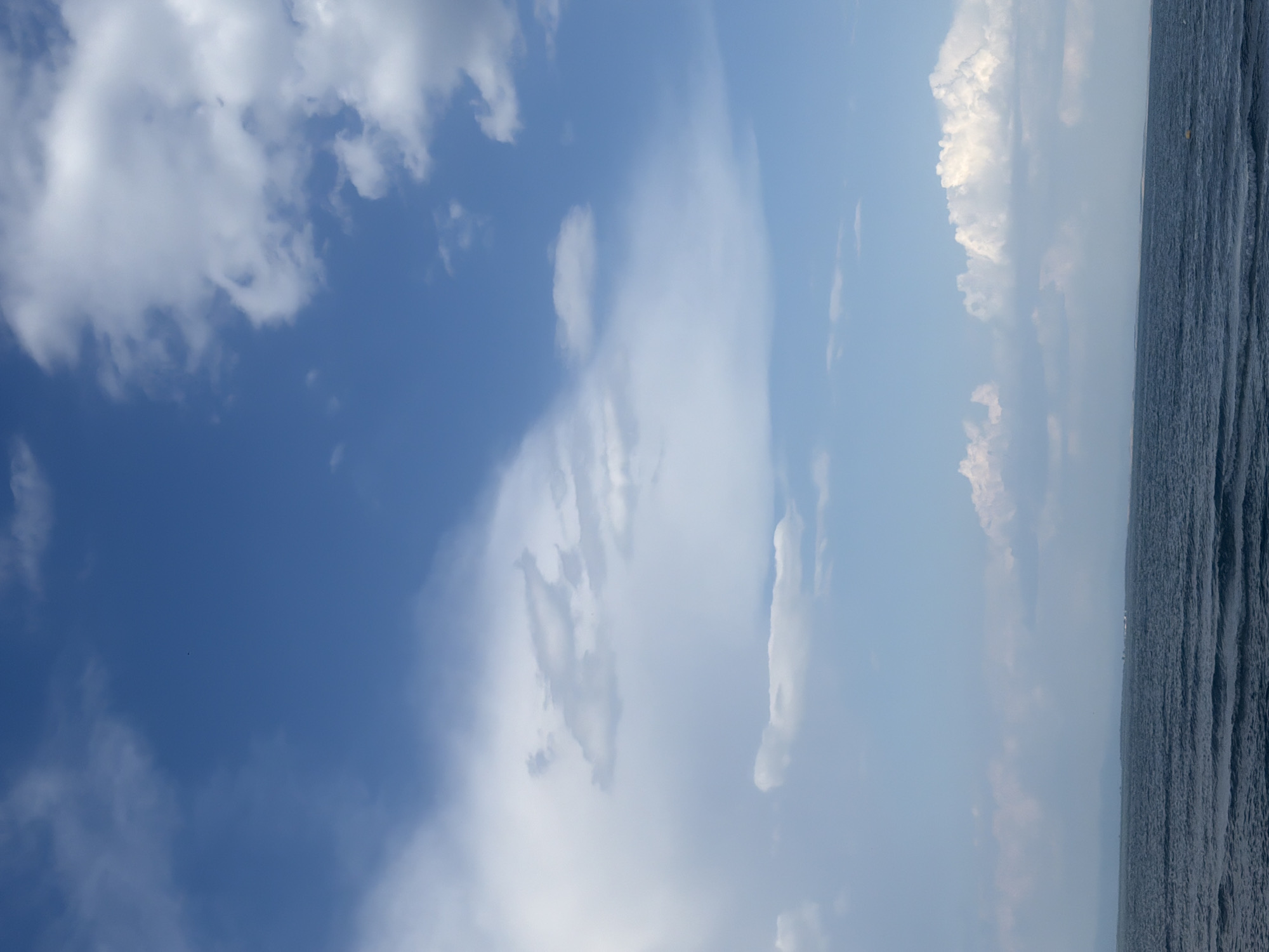 This is a vertical, wide-angle photograph featuring a dramatic sky and a body of water. The sky dominates the left side of the frame, transitioning from a deep blue at the top to a lighter, hazy blue towards the horizon. It is filled with a variety of clouds. On the left, large, puffy white clouds are prominent, while towards the center and right, there are wispy, translucent clouds that appear to be thinner and more spread out. The right side of the image shows a dark, textured body of water, likely the sea, with a slightly undulating surface that creates a ribbed pattern. A faint line of land or distant structures is visible on the far right horizon, barely discernible through the haze.
The image appears to be taken during the daytime, possibly late afternoon or early evening, given the soft lighting and hazy atmosphere. The weather seems to be clear with scattered clouds. The scene evokes a sense of vastness and tranquility, with the expansive sky and the calm water. There are no visible people or objects of interest in the foreground. The composition suggests a serene coastal or oceanic landscape. There is no visible text in the image.