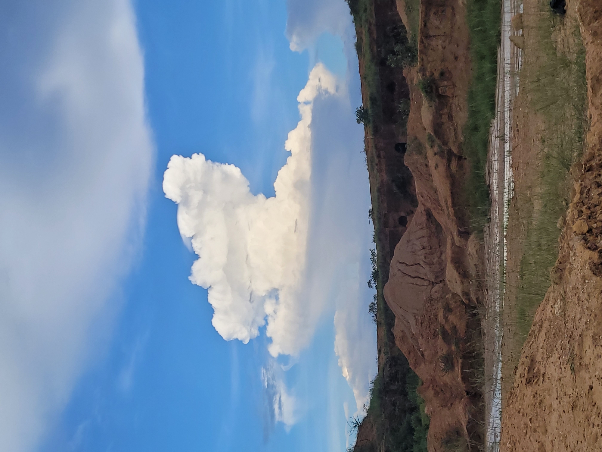 A wide-angle, landscape photograph captures a partially excavated hillside under a blue sky dotted with fluffy white clouds. The foreground is dominated by mounds of reddish-brown soil, some with sparse green grass. A shallow, muddy stream or pool of water runs through the middle ground, reflecting the sky. The hillside itself shows evidence of excavation, with exposed earth and some dark openings, possibly caves or tunnels. Green vegetation grows along the top of the hillside and in patches on its slopes. The overall scene suggests an active or abandoned quarry or construction site in an arid or semi-arid environment. The image is likely taken in Bukuru, Nigeria, given the geographical context.