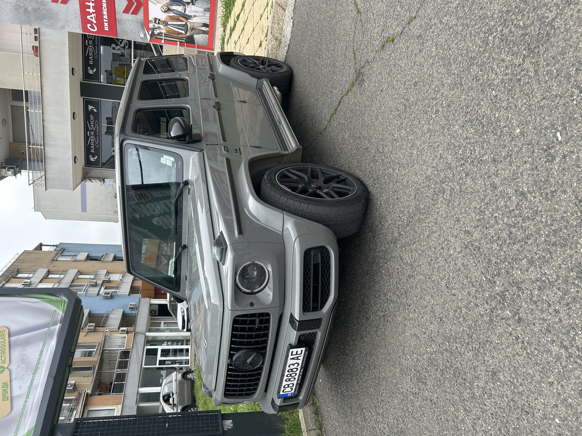 A grey Mercedes-Benz G-Wagen (G63 AMG model) is parked on an asphalt street, visible from a frontal-left perspective. The luxury SUV features black multi-spoke alloy wheels with red brake calipers, and a "V8 BITURBO" badge on its side fender. The front grille prominently displays the Mercedes-Benz star logo, and the Bulgarian license plate reads "CB 8883 AE," indicating its origin from Burgas, with "SILVER STAR" printed below.
In the background, a two-story building features black and white signs for "BARBER SHOP ROYAL SCISSORS" and "BARBERS ROYAL SCISSOR." Adjacent to it, a red sign is partially visible with Cyrillic text including "САН" and "КИТАЙСКИ" (Chinese). Further along the street, a multi-story residential or commercial building is visible with numerous windows and air conditioning units. On the far left, a large billboard for "ACTIREGULARIS" and "ДРОЖДИ" (yeast) is partially obscured, referencing the Activia brand. Another shop sign reads "Forelle" and "КРЕДИТИ" (Credits) with a phone number "0876 872 266."
The scene is an urban streetscape, likely in Burgas, Bulgaria, as evidenced by the license plate and the widespread use of Cyrillic text on signs. The asphalt road shows minor cracks and sparse patches of grass. The time of day appears to be daytime, under an overcast sky, providing diffused natural light. No individuals are visible in the image.