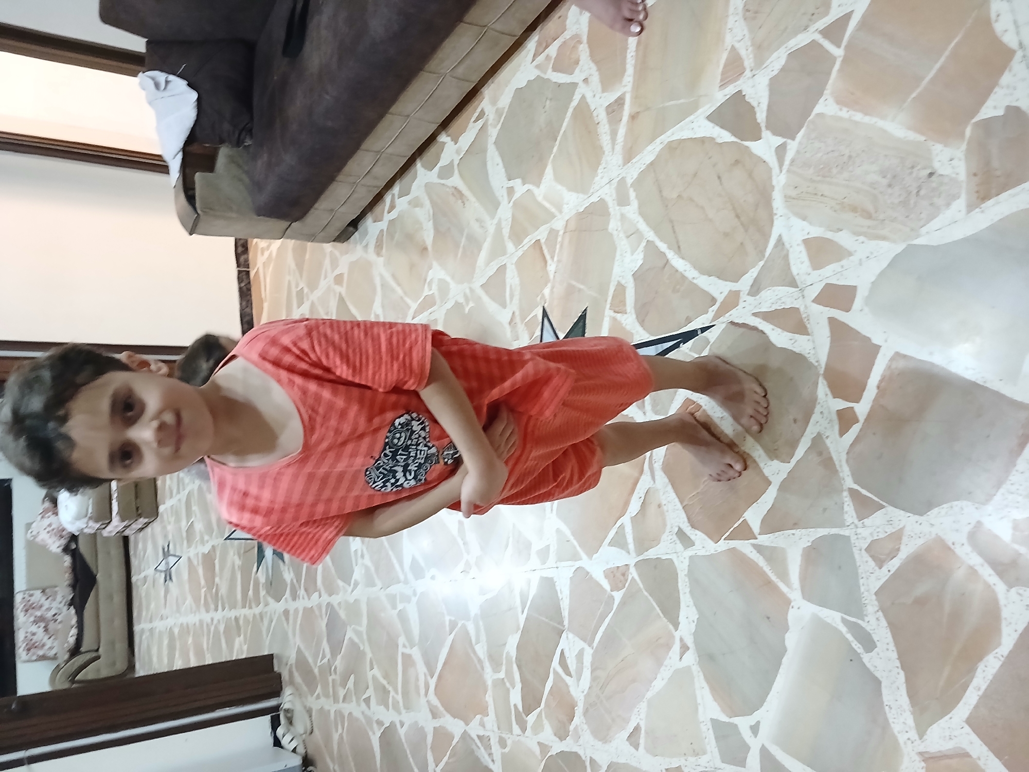 A young boy stands on a marble-patterned floor in what appears to be a living room. He is wearing a coral-colored t-shirt with a graphic design and matching shorts. His hands are clasped in front of him. In the background, a couch with dark brown cushions and a lighter brown base is visible. A white cloth is draped over the armrest of the couch. To the left, a portion of a doorway with a dark brown frame is seen, along with the corner of a cream-colored wall. The floor is tiled with irregularly shaped pieces of light brown, beige, and white marble, separated by white grout lines. The overall environment suggests an indoor setting, likely a home.