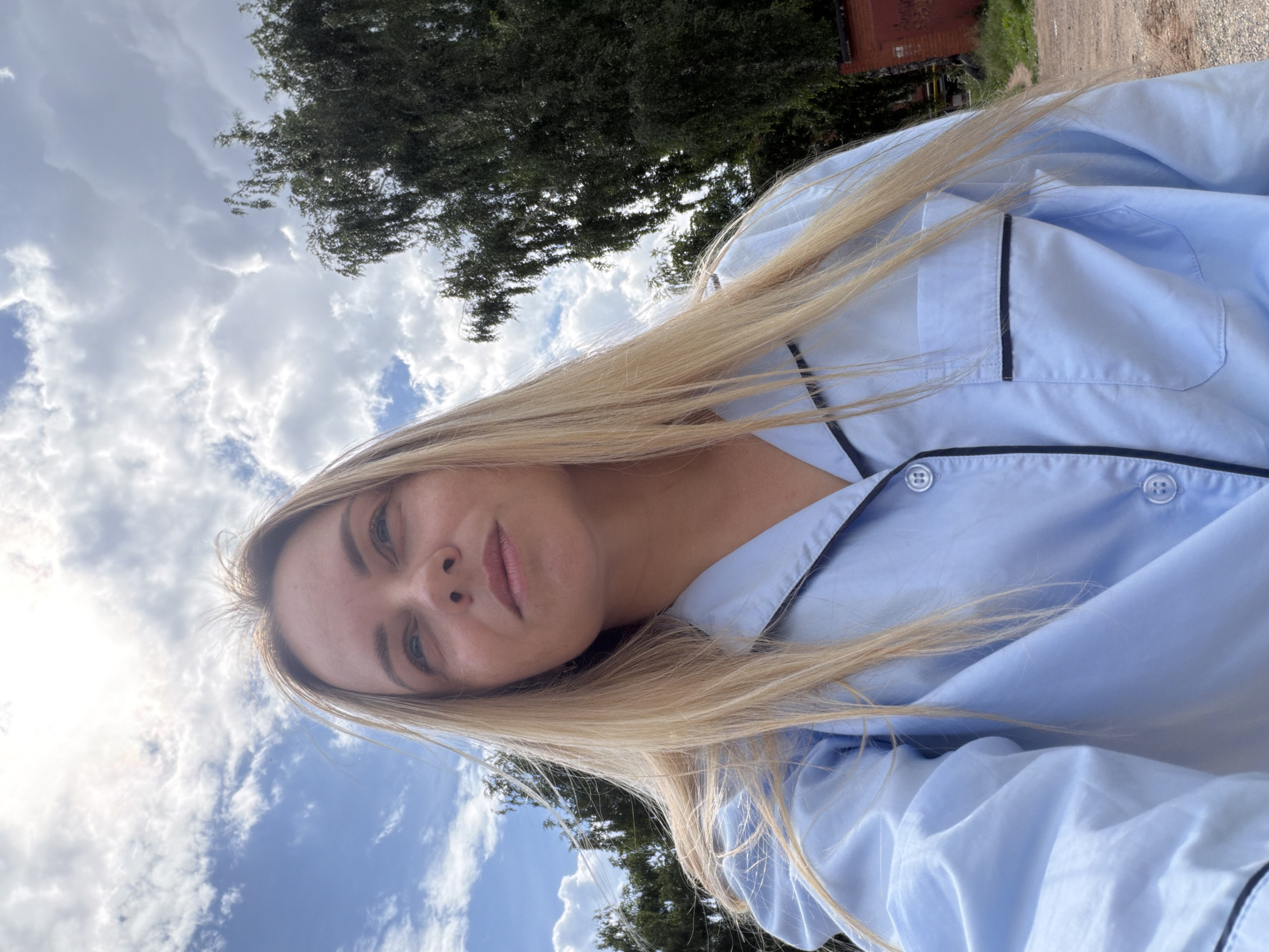 A young blonde woman is captured in a low-angle, outdoor selfie, looking upwards towards a cloudy sky. She is wearing a light blue pajama top with black trim and buttons. Her hair is long and straight, falling around her shoulders. The background consists of a sky with scattered white clouds and the sun partially obscured, along with the lush green foliage of trees and a glimpse of a red brick building. The ground appears to be dirt or gravel, suggesting a rural or suburban setting. This image was taken in Votkinsk, Russia.