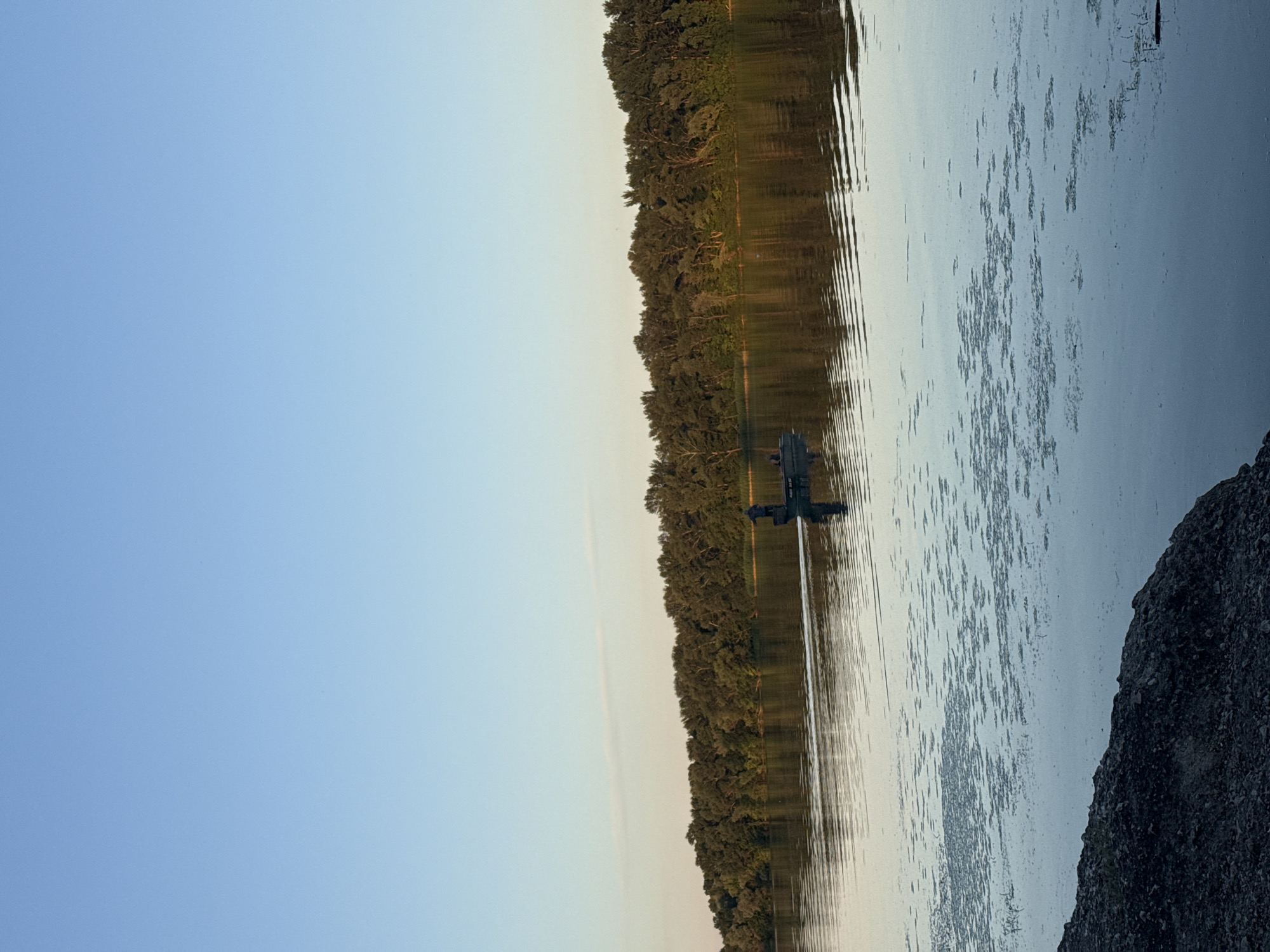 The image depicts a serene natural landscape with a clear blue sky transitioning to a pale yellow hue near the horizon. A dense line of trees forms a dark green barrier along the far bank of a wide river. The river's surface is calm, reflecting the sky and trees, with small patches of aquatic vegetation visible. In the mid-ground, a small boat with at least one person standing is moving across the water, leaving a wake. The foreground features a rocky shoreline. This scene is likely located in the city of Dnipro, Ukraine, possibly along the Dnipro River.