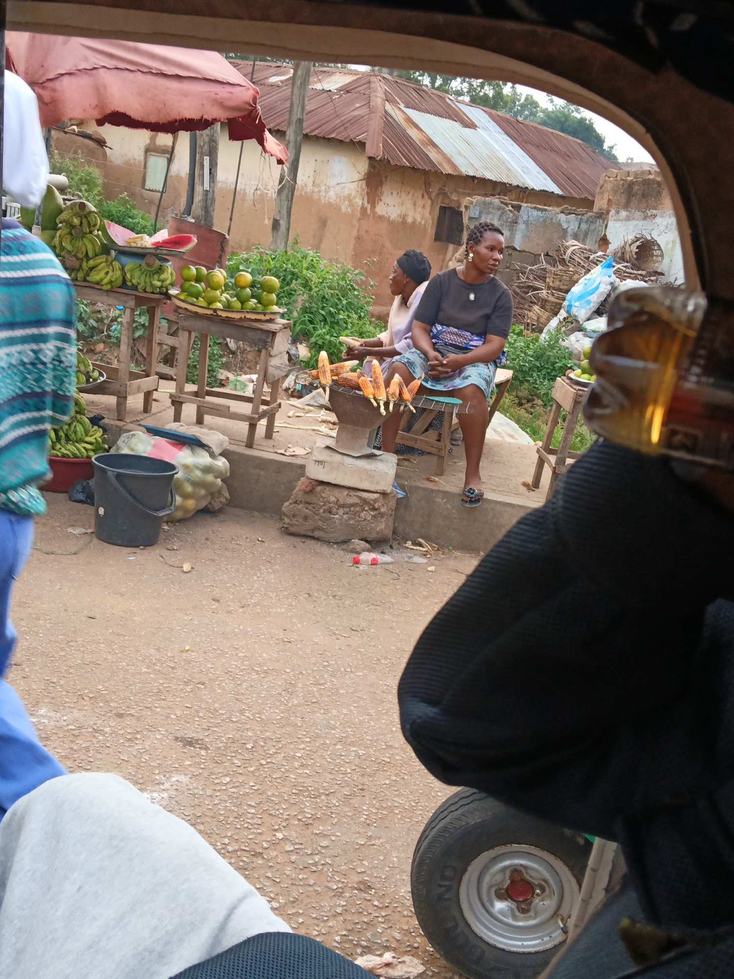 The image shows an outdoor scene, likely a market, with two women in the background. One woman, wearing a dark shirt and a patterned skirt, is seated on a stool, facing left. Her hands are clasped in her lap. The other woman, wearing a light-colored garment and a dark head covering, is seated to her left, also facing left. In front of them, on a wooden stand, are several ears of corn being roasted. To the left, a variety of fruits, including bananas and oranges, are displayed on wooden tables. A black bucket and a bag of produce are on the ground. The background features buildings with corrugated metal roofs and mud-colored walls. The foreground is dominated by the interior of a vehicle, with a dark fabric covering the right side and a tire visible on the bottom right. The ground is dusty and uneven.
