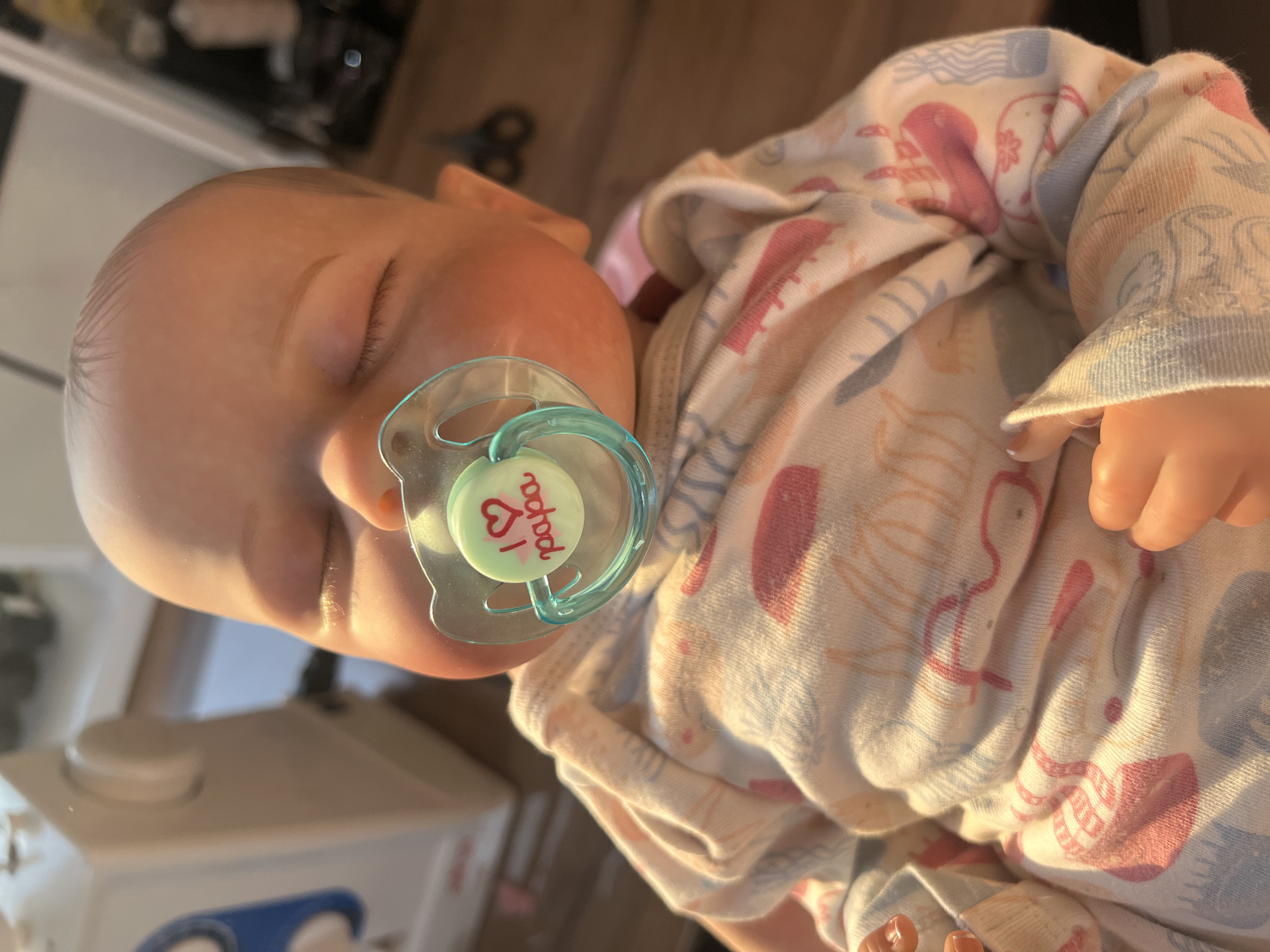 This is a close-up, high-angle, vertical shot of a baby doll lying down, facing left. The doll is wearing a light-colored onesie with a pattern of pink and blue sea creatures and abstract shapes. A teal pacifier is in the doll's mouth. The pacifier has a green disc with red text that reads "I love papa" and a small red heart icon. The doll's eyes are closed, and its skin appears realistic, with visible eyelashes. The background shows a wooden surface and what appears to be a white appliance, possibly a sewing machine. The lighting suggests it is daytime, with sunlight casting a warm glow on the doll. The overall impression is one of peacefulness and affection.