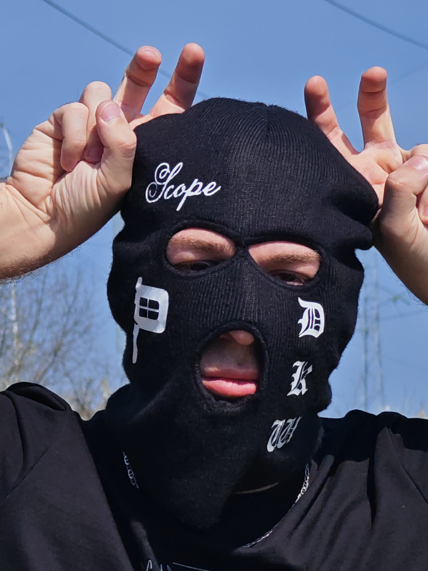 A close-up portrait depicts an individual outdoors, framed from the chest up, under a clear blue sky. The person is wearing a black balaclava that completely covers their face, featuring three cutouts for the eyes and mouth. Visible through the eyeholes are the individual's brown eyes, while the mouth cutout reveals a slightly open mouth with the tongue partially extended.
The balaclava is adorned with white embroidered elements: "Scope" in a cursive font is positioned above the left eye cutout. Below the left eye cutout, a stylized letter "P" with an embedded window graphic is present. On the right side of the face, a stylized letter "D" is visible below the right eye cutout, and a stylized letter "K" is situated below it. Further down, beneath the mouth cutout, a stylized letter "W" is visible.
The individual's hands are raised to either side of their head, just above the balaclava. Both hands exhibit an identical gesture: the index and middle fingers are extended upwards, while the thumb and remaining fingers are curled, forming a "bunny ears" or inverted V-sign. The person is wearing a black garment, possibly a t-shirt or hooded top, and a silver-colored chain is visible around their neck, partially obscured by the balaclava and clothing.
The background consists primarily of a clear, bright blue sky. Sparse, bare tree branches are discernible on the lower left and right sides of the frame, indicating a wooded area without foliage. A thin, dark line, consistent with a power line or cable, stretches across the upper background. Faint, distant structures, possibly power poles, are visible on the right side of the background. The specific location within the city of Shumen, Bulgaria, is not discernible from the visual information provided.