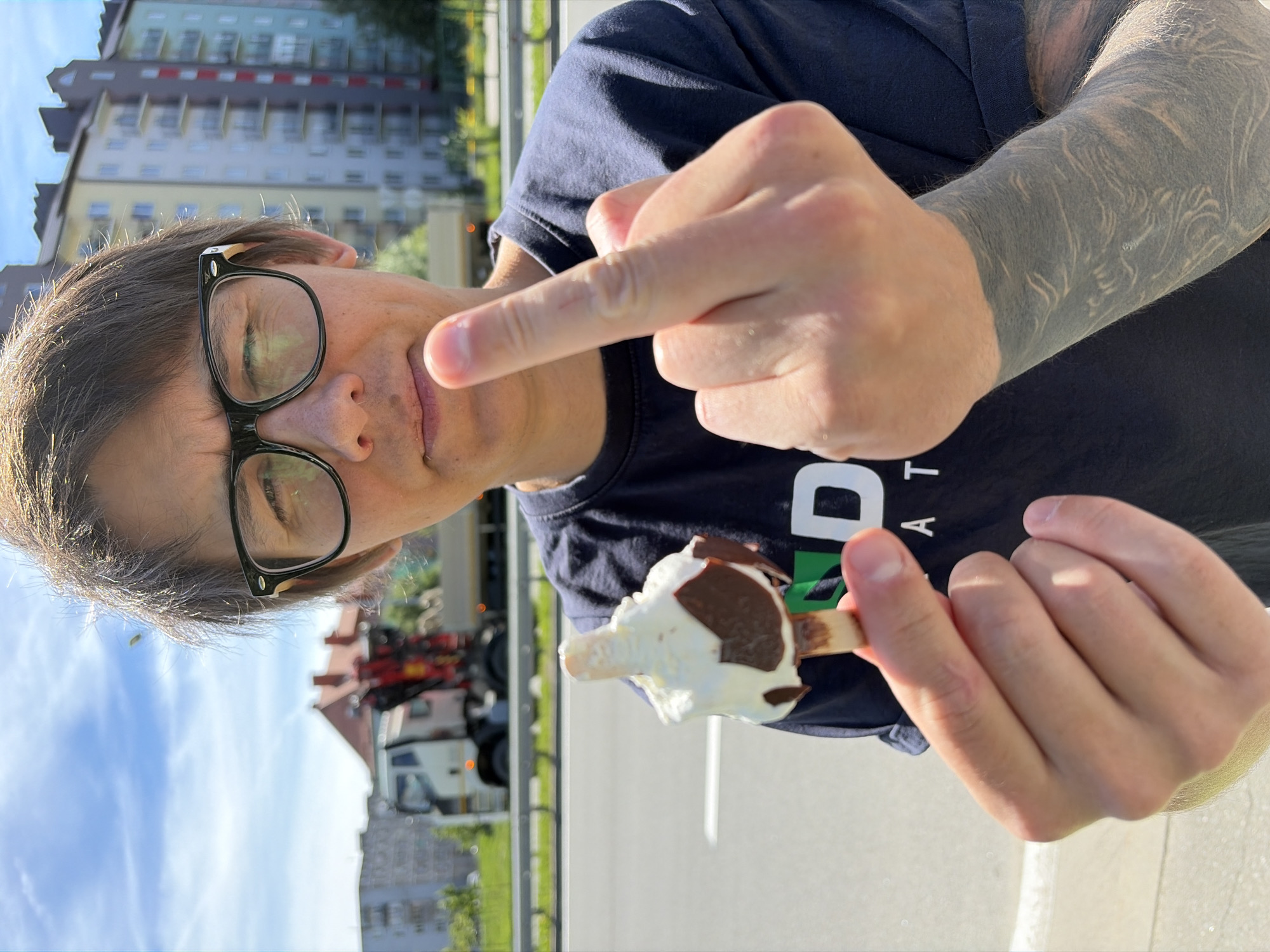 The media file is a slightly rotated, medium close-up shot of a person outdoors, holding a melting popsicle. The person, who appears to be wearing glasses and a dark t-shirt, is gesturing with their right hand. Their facial expression is not clearly discernible due to the angle and the focus on their hand and the popsicle. The popsicle itself is white with streaks of chocolate and is beginning to melt.
In the background, there are buildings that appear to be apartment complexes, with a clear blue sky overhead, suggesting it is daytime. The presence of a road and what looks like a construction vehicle or truck indicates an urban or suburban environment. The lighting suggests a sunny day. There is a visible tattoo on the person's left arm, which appears to be a large, detailed design. The t-shirt has some white text on it, but it is partially obscured and not fully legible. The overall mood is casual and perhaps a bit defiant due to the gesture.