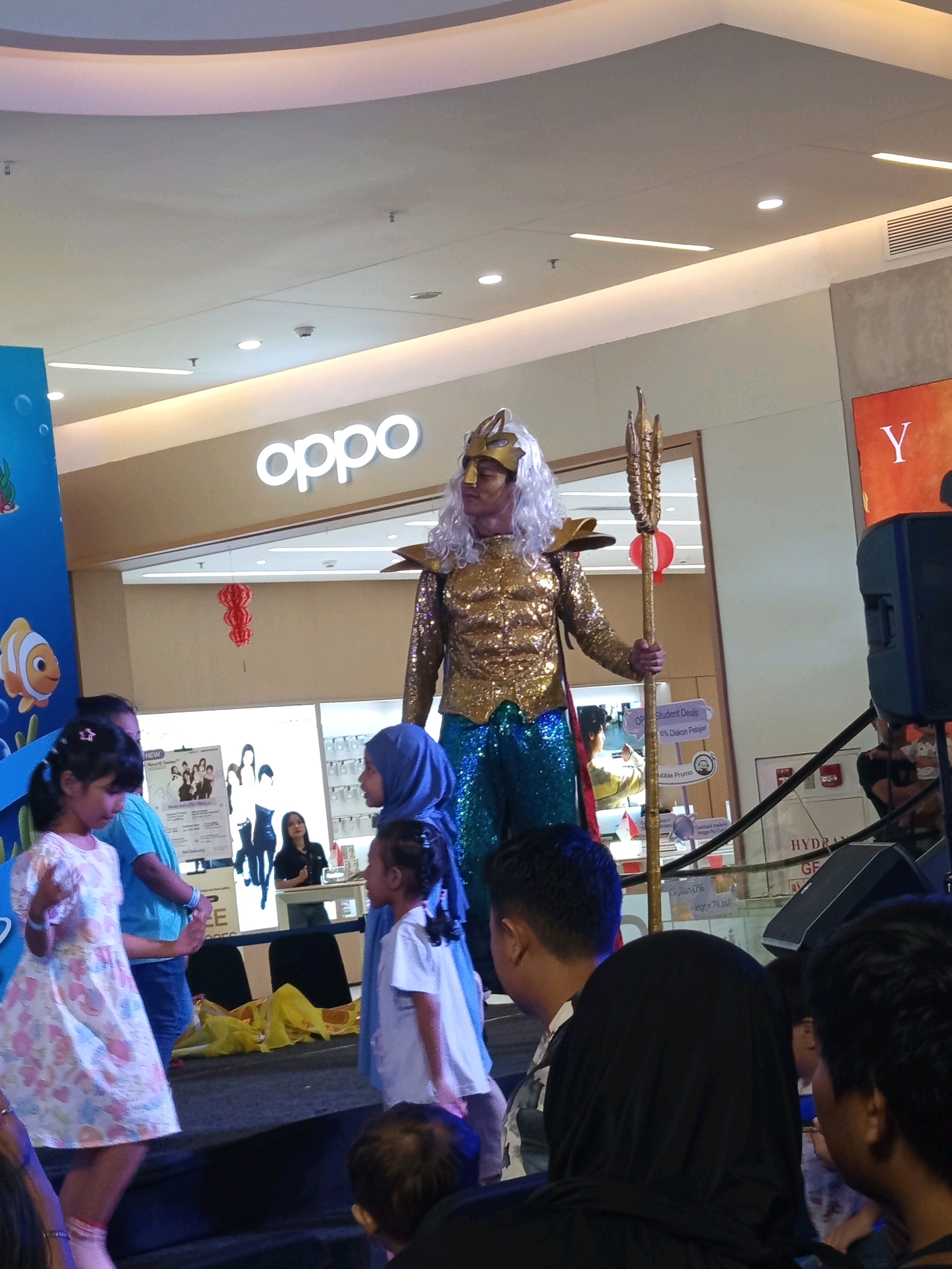 An event is taking place at a shopping mall in Ciakar, Indonesia, likely at the Lippo Plaza. A man dressed as Aquaman, with a gold sequined top and blue sequined pants, stands on a stage holding a trident. He has long blonde hair and a gold crown. Several children are on the stage with him, including a young girl in a floral dress and another in a white t-shirt and blue headscarf. Adults are also present, some watching the performance, others participating. A blue banner with a cartoon fish is visible on the left side of the stage. Behind the stage, there is a store with an "OPPO" logo and promotional materials. The ceiling of the mall, with its lighting and air conditioning vents, is visible overhead.