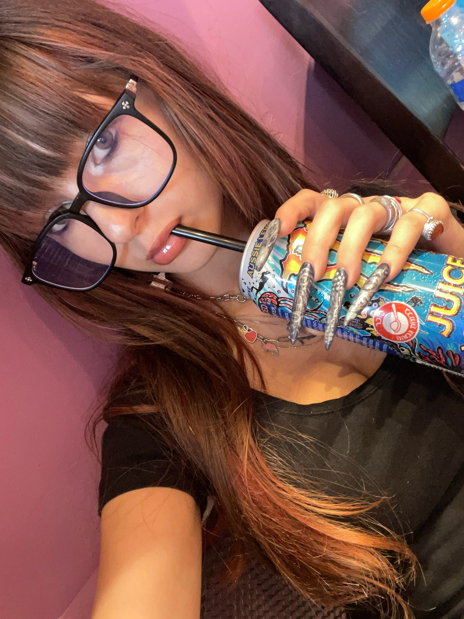 A close-up, eye-level, slightly angled medium shot shows a young woman with long, layered brown hair with reddish highlights, and bangs, wearing black-rimmed glasses with small cross charms on the temples. She is holding a dark straw to her lips, which are painted with glossy pink lipstick, and is drinking from a can of "Aussie Juice" with colorful graphics. Her hands, adorned with multiple silver rings and long, grey, pointed nail extensions, grip the can. She is wearing a black t-shirt, and a delicate silver necklace with a red heart charm is visible around her neck. The background consists of a pink wall on the left and a dark surface on the right, with a partially visible plastic water bottle with an orange cap. The image is taken indoors, likely in a casual setting. There is no specific indication of the location being in Raanana, Israel.