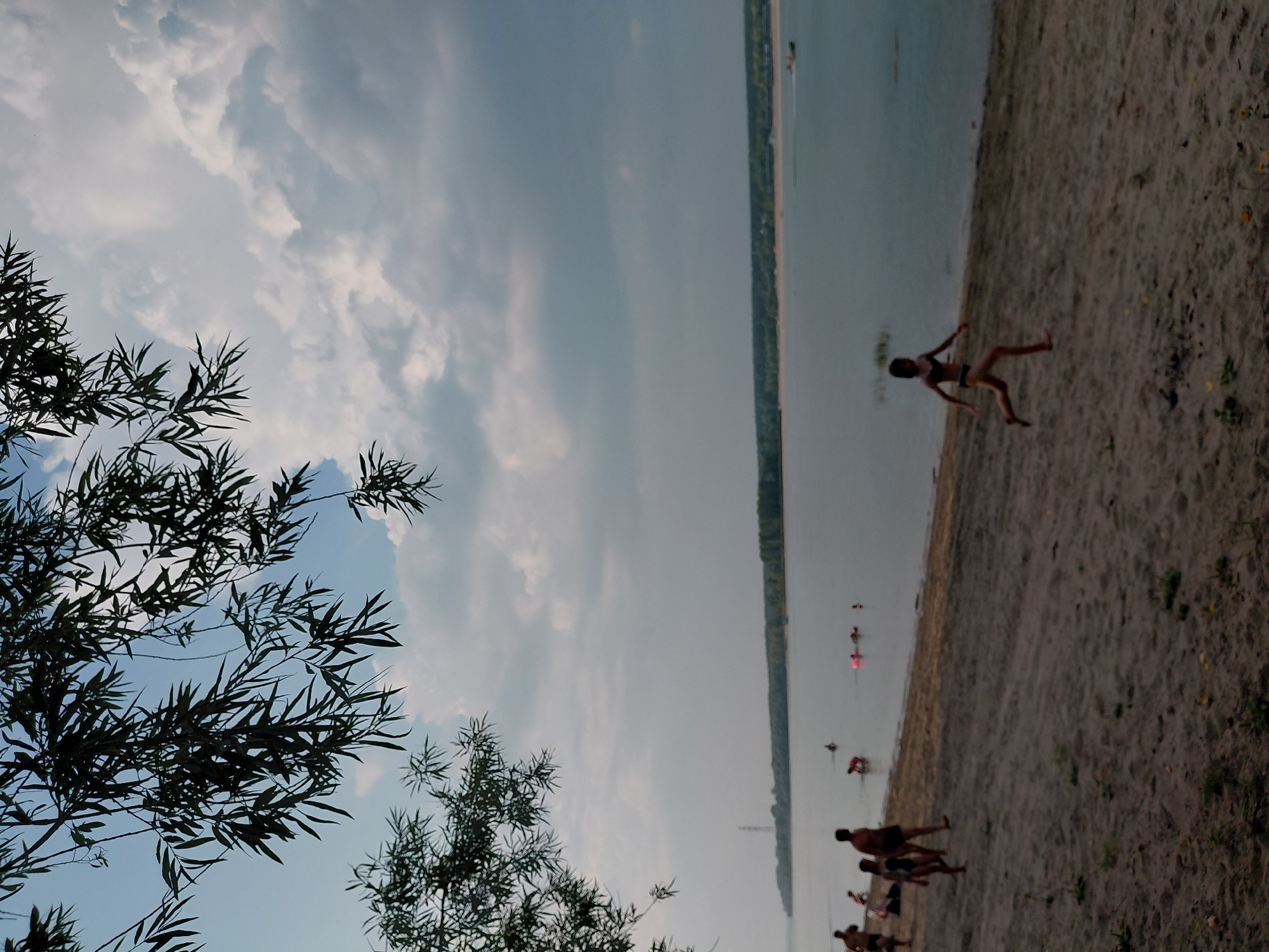The image depicts a sandy beach adjacent to a wide river. In the foreground, on the beach, a young girl in a dark swimsuit is running towards the right. To the left of the running girl, several other people, including adults and children, are visible on the beach. Some are walking, while others are near the water's edge. In the river, a few individuals are swimming or wading. The water is calm, and a distant treeline marks the opposite bank of the river. The sky is partly cloudy, with dramatic cloud formations. The scene suggests a recreational area, possibly a popular spot for swimming and leisure. Given the context of Novosibirsk, Russia, this location could be a beach along the Ob River, a prominent waterway flowing through the city.