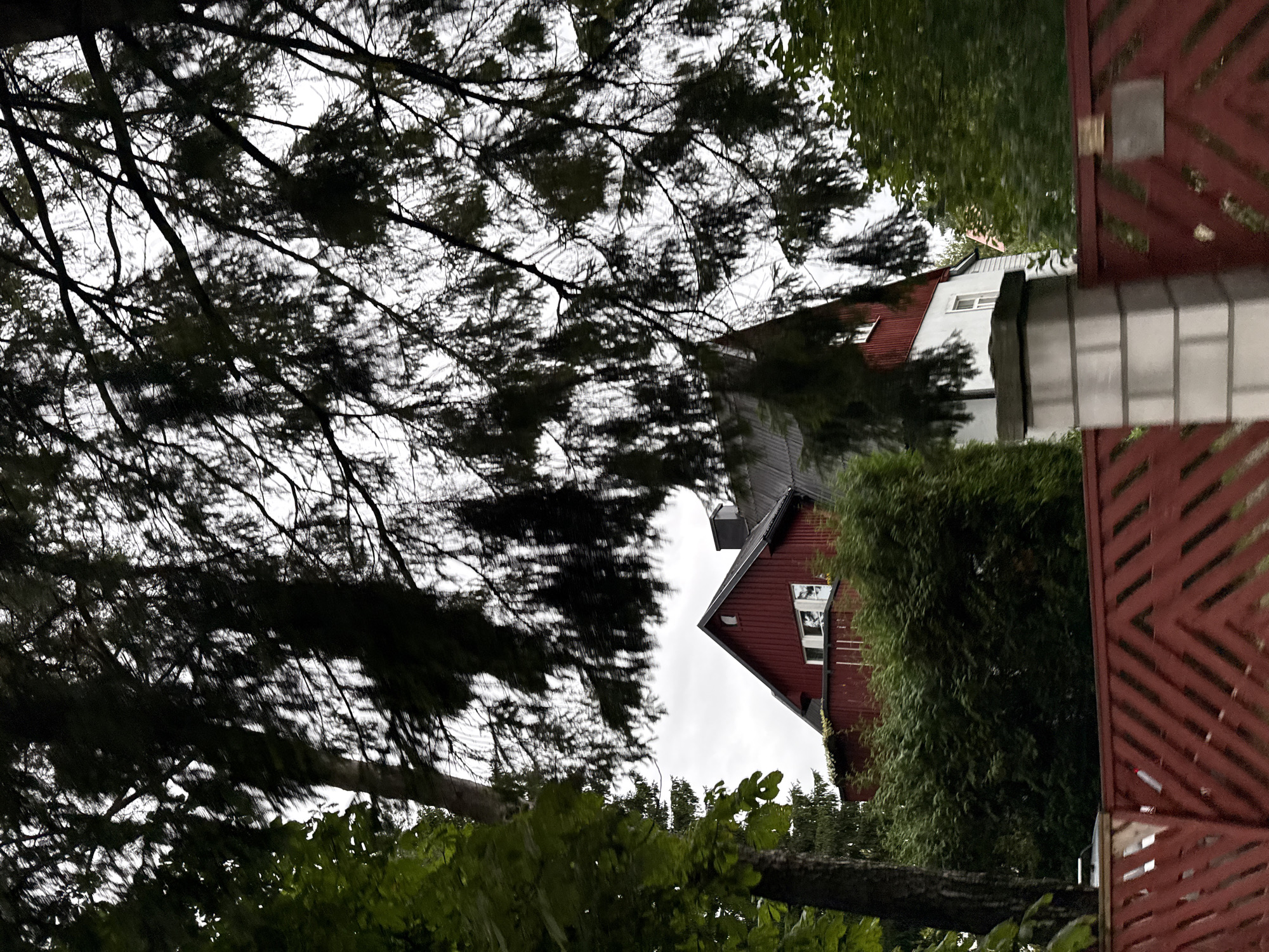 A dark, overcast sky looms above a two-story red and white house, partially obscured by the dense branches of a coniferous tree. The house features a dark gray roof and a chimney. In the foreground, a vibrant red wooden fence runs horizontally across the image, with a white brick pillar to its right. Lush green foliage surrounds the house and fence, suggesting a suburban or residential area within the city of Tallinn, Estonia. The overall scene conveys a sense of privacy and tranquility, with the natural elements framing the architectural structures.