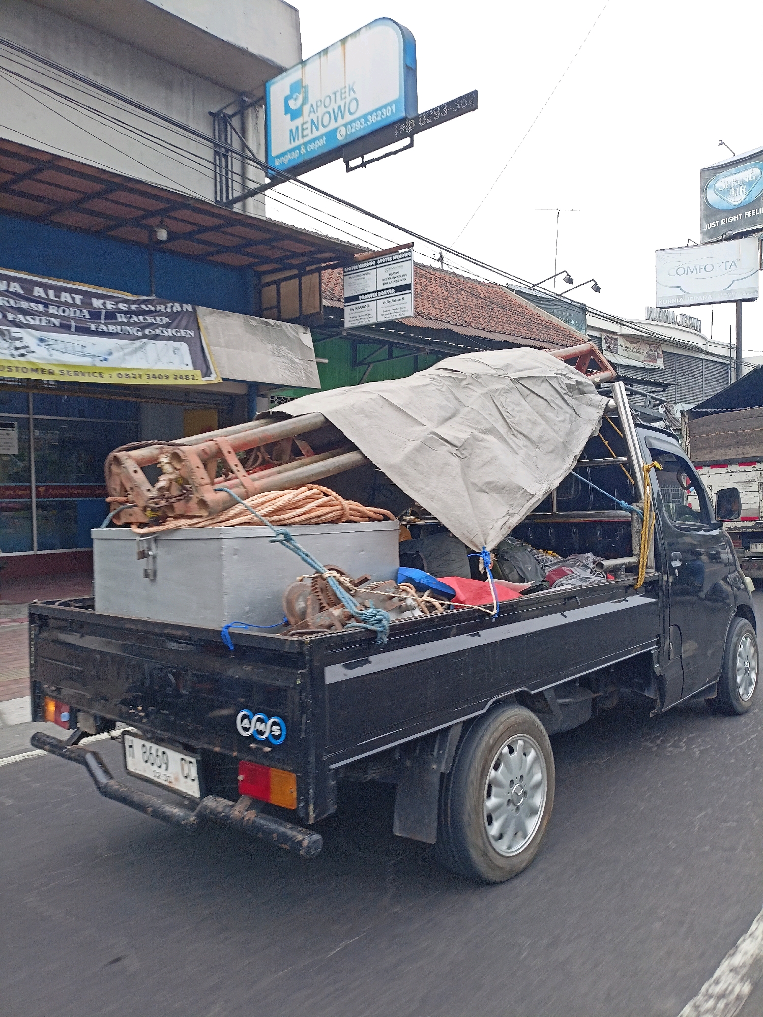 A black pickup truck, loaded with equipment including a large metal frame covered by a tarp and a gray toolbox, is driving on a street in Magelang, Indonesia. The truck has a license plate reading "H 8669 DD". The truck is in motion, as indicated by the motion blur. In the background, there are several signs, including one for "APOTEK MENOWO", another for "OMFORA", and a smaller one that says "SHINE AIR". The street is paved and appears to be a commercial area with buildings lining the side. The sky is overcast, suggesting cloudy weather.