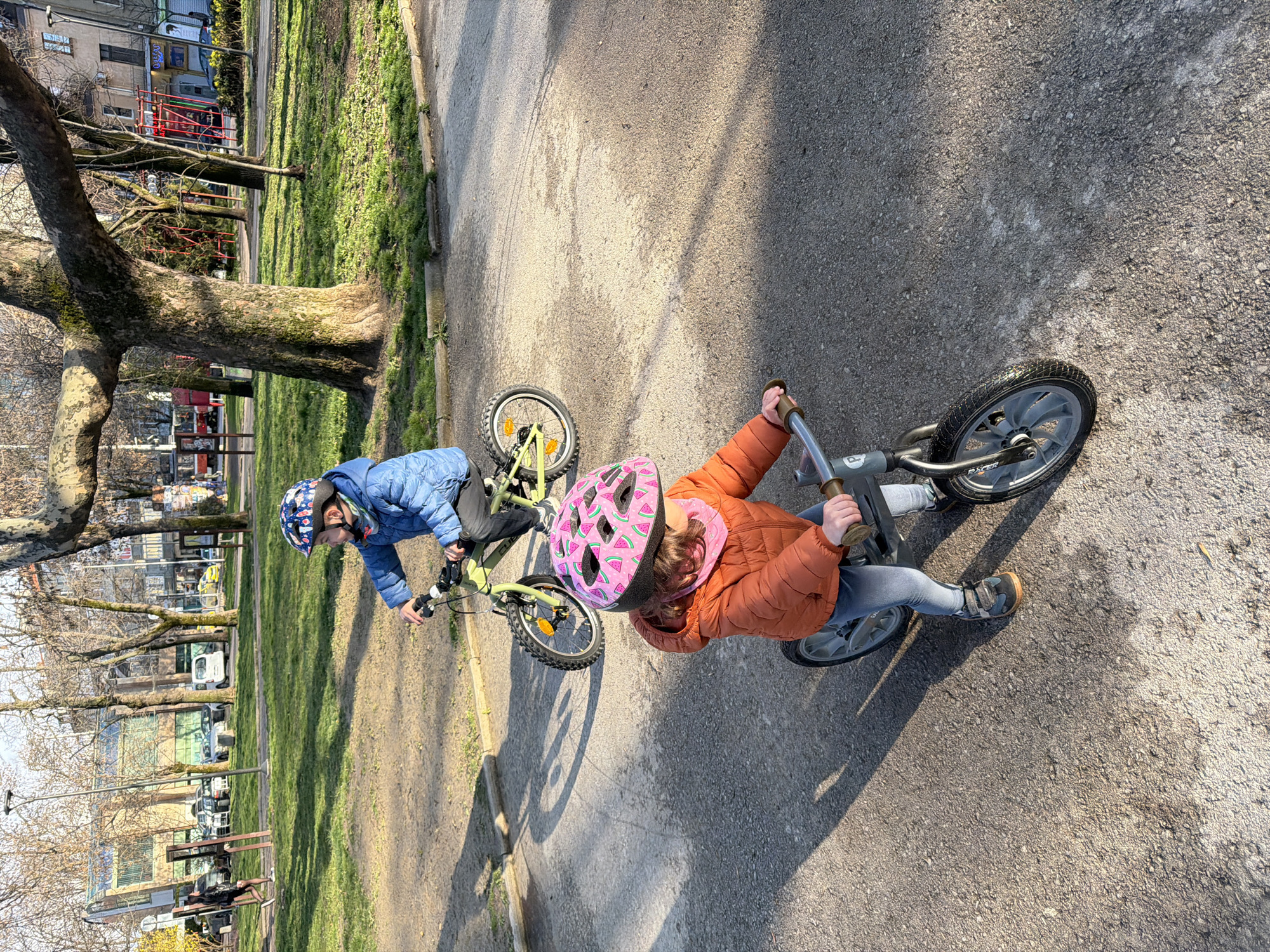 Two children are riding bikes on a paved path in Doktorska Gradina (Doctor's Garden), Sofia, Bulgaria, illuminated by sunlight that casts long shadows. In the foreground, a young child, approximately 3-5 years old, wears an orange puffer jacket, light blue jeans, and a pink helmet with a watermelon pattern over a pink neck warmer, operating a grey balance bike with their feet. Behind and to the left, an older child, approximately 5-7 years old, wears a blue puffer jacket, dark trousers, and a blue patterned helmet, riding a lime-green pedal bicycle. Both children are positioned as if moving towards the viewer and slightly to the right. The paved path is bordered by sparse green grass and bare deciduous trees. The background reveals an urban setting with various buildings, including the distinct modern, green-roofed "Red House Centre for Culture and Debate" on the left, parked cars, a red tram, and red playground equipment on the right. A solitary individual in dark attire walks on a path in the distant left background.