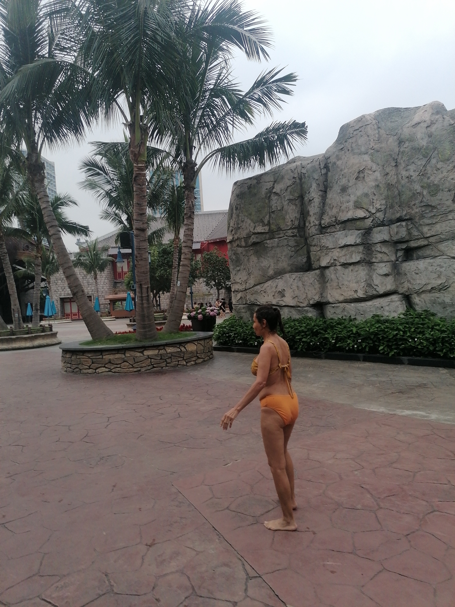 The image depicts an outdoor scene, likely a resort or leisure area, consistent with locations found in Bai Chay Ward, Vietnam. In the foreground, a woman is walking barefoot from left to right across a reddish-brown, patterned paved surface. She is seen from a side-rear perspective, wearing an orange two-piece swimsuit, with her hair pulled back. Her relaxed posture suggests a casual stroll.
The background is rich with tropical and landscaped features. Several tall palm trees with green fronds are prominent, some planted within a low, stone-faced circular planter that also contains grass and red flowers. Behind the palm trees on the left, glimpses of buildings with red roofs and traditional-style architecture are visible, alongside small blue umbrellas that suggest outdoor seating. Further in the distance, taller, modern high-rise buildings peek through the foliage.
To the right of the woman, a large, grey, textured rock formation serves as a significant backdrop element, likely an artificial feature designed for landscaping. A low, dense hedge of green bushes runs along the base of this rock wall. The sky is overcast and white, indicating a cloudy daytime, and there is no direct sunlight visible. The overall setting conveys a relaxed, tropical, and recreational environment. No visible text is present in the image.
