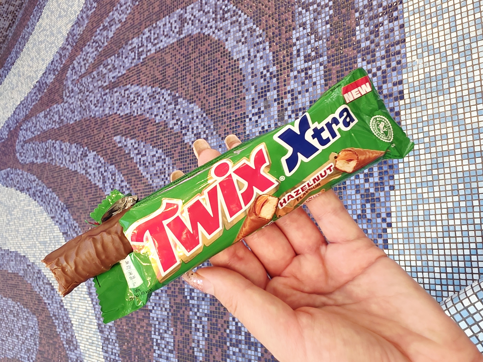 The media file shows a person's hand holding a Twix Xtra chocolate bar. The chocolate bar is green with "Twix Xtra" written in large red and blue letters. A portion of the chocolate bar is unwrapped, revealing the chocolate coating and cookie base. The background appears to be a mosaic tile surface with a pattern of purple, white, and brown. The lighting suggests it might be daytime, but the specific time of day and weather are not determinable. The person's nails are decorated with a light pink glitter polish. Text visible on the packaging includes "NEW" and "HAZELNUT FLAVOUR". The overall scene appears to be a casual presentation of the candy bar.
