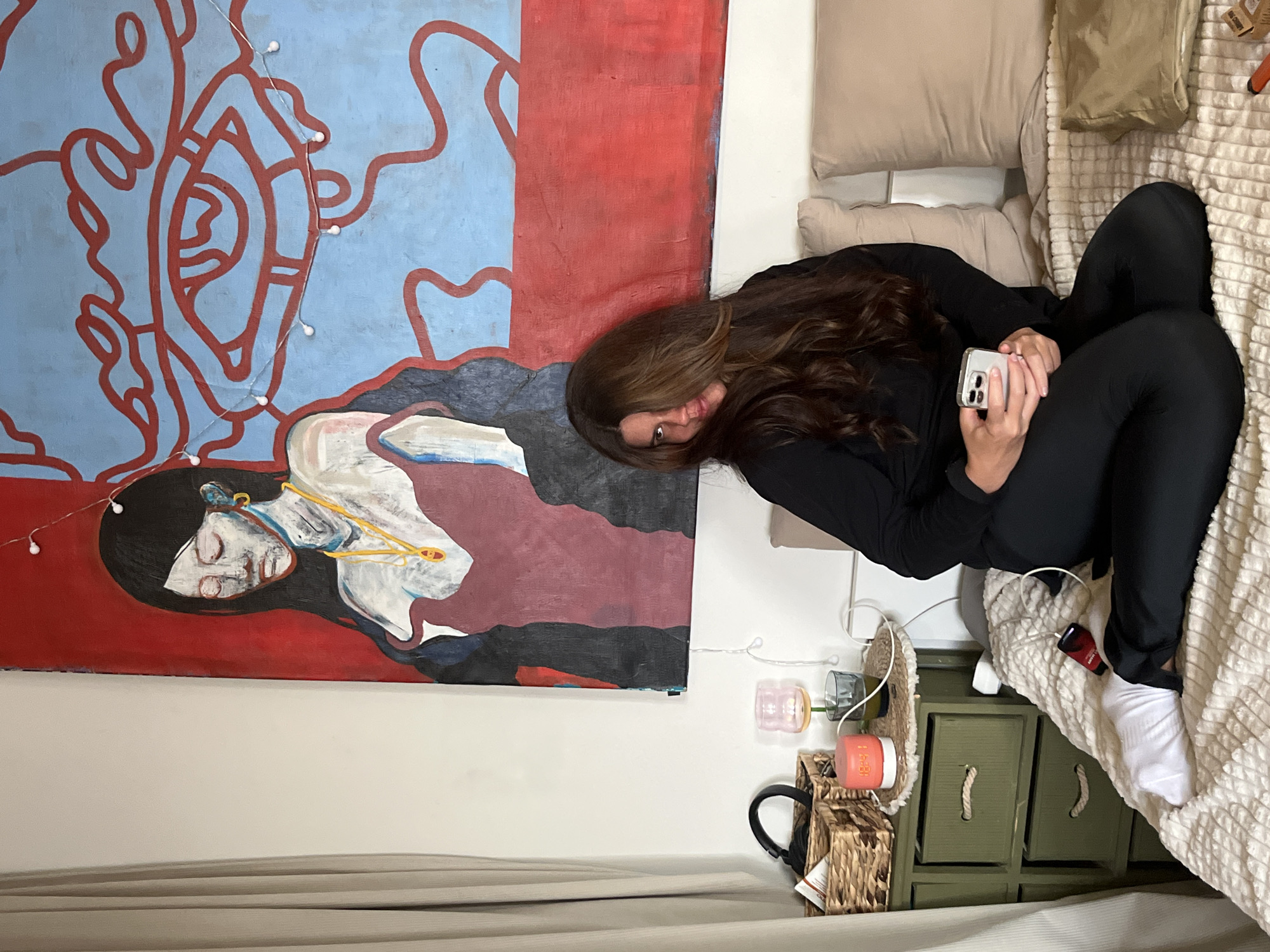 A woman with long brown hair lies on a bed with off-white bedding and pillows. She is wearing black clothing and white socks, and she is holding a smartphone in her hands. To the left of the woman is a large painting on the wall. The painting has two distinct sections: a blue section with red abstract lines and a red section with a woman's face, neck and part of her torso depicted. String lights are attached to the painting. Next to the bed is a small, green cabinet with wicker baskets and other small objects on top. There is also a curtain visible at the bottom of the image. There is no evidence of Krasnoyarsk, Russia.