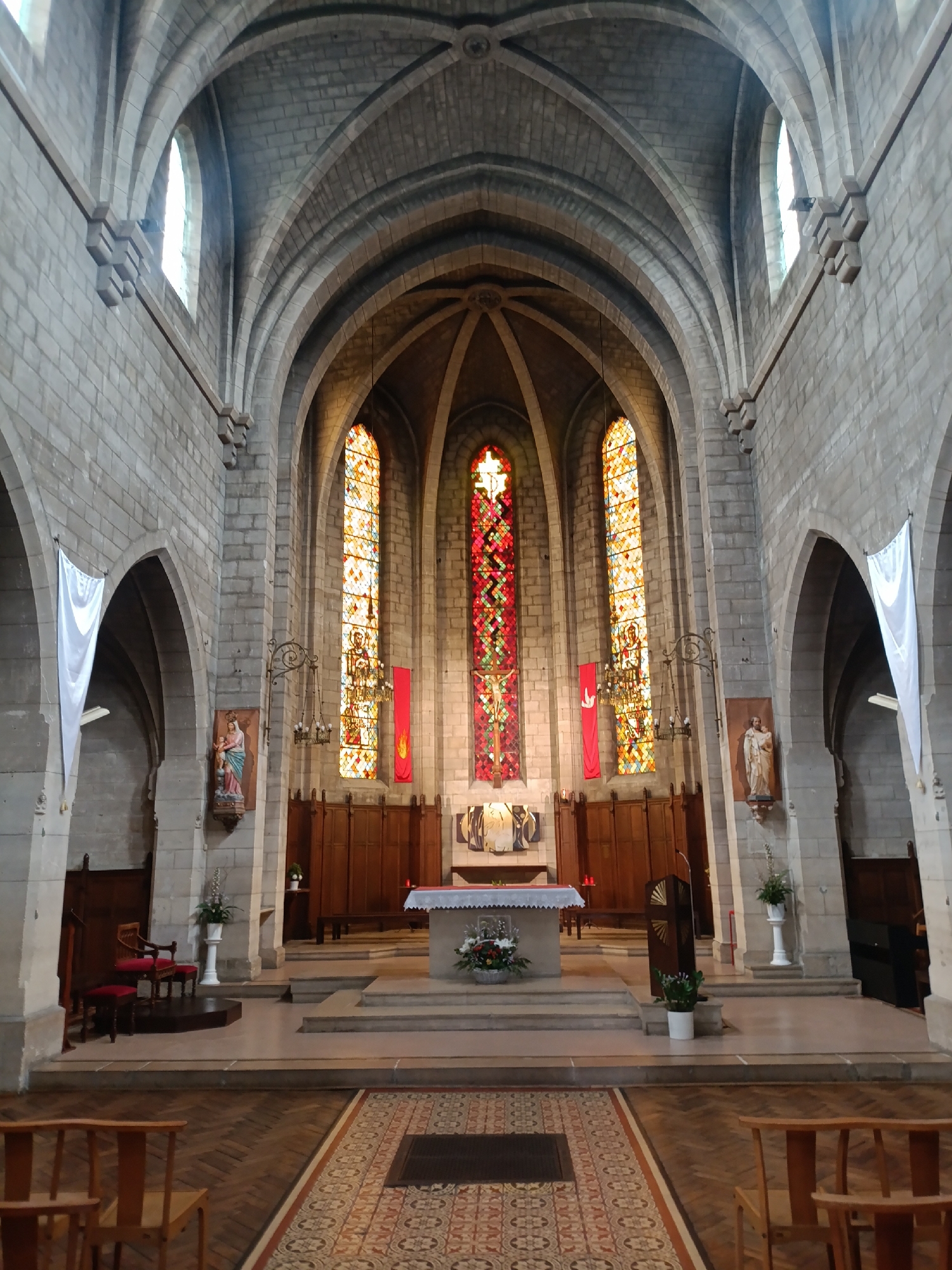 The image shows the interior of a church. The architecture is Gothic, with stone walls and arched ceilings. The focal point of the image is the altar area, which is elevated and features a white altar cloth adorned with flowers and a cross. Behind the altar, there is a wooden choir stall and a large stained-glass window with a red and white design. To the left of the altar, there is a pulpit.
On either side of the altar, there are alcoves with statues and stained-glass windows depicting religious figures. White banners hang from the walls on the sides. In the foreground, there are wooden pews and a patterned rug. The floor is made of wood. The lighting is dim, with natural light entering through the stained-glass windows. The overall impression is one of solemnity and religious devotion.