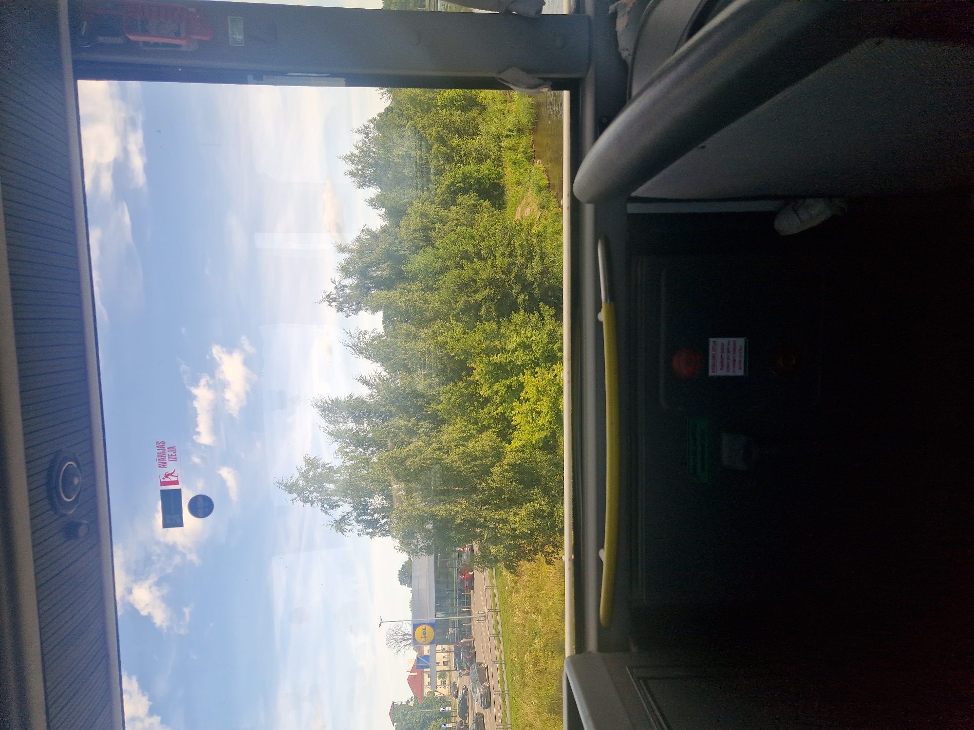 The image shows a view from inside a bus, looking out through the windshield. Outside, a bright blue sky with scattered white clouds is visible. In the distance, there are green trees, a body of water, a road with cars, and a building with a LIDL logo. There is a sign on the windshield that reads "AVĀRIJAS IZEJA" in red and white lettering. The interior of the bus is dark, with some yellow and gray plastic elements visible. There are also some buttons and signs on the interior panels, including a green sign and a red emergency button. The bus appears to be stopped, possibly at a bus stop or a traffic light, given the parked cars and the LIDL supermarket in the background. The environment suggests an urban or suburban setting in Ogre, Latvia, as indicated by the LIDL store and the Latvian text on the sign.