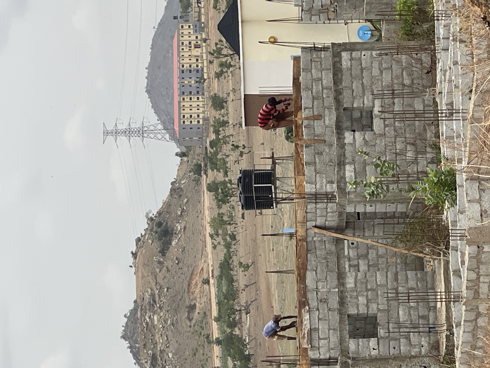 The image captures a construction site in Angwan Sayawa, Nigeria. Two individuals are working on the upper floors of a multi-story building made of concrete blocks and reinforced with rebar. One worker, in a red and black striped shirt, appears to be working on the higher level. The second worker, in a light-colored shirt, is on a lower level. A black water tank is visible on the site. In the background, a mountainous landscape is present with sparse vegetation. A high-voltage power line tower is also visible. A multi-story building stands in the distance. The sky is overcast.