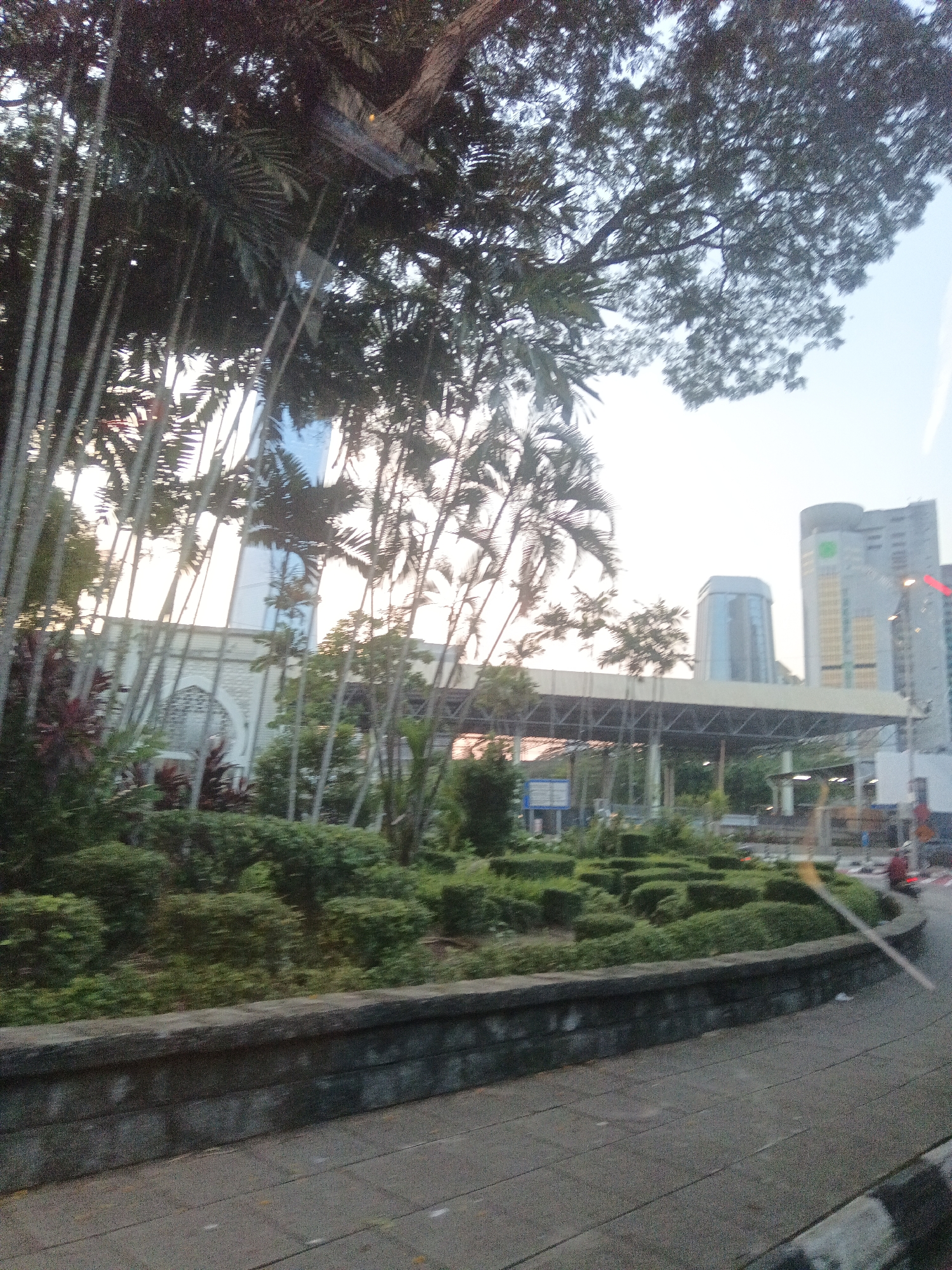 The image shows a view of a city street in Kuala Lumpur, Malaysia, with lush greenery and modern architecture. In the foreground, there is a stone wall and a paved walkway. Behind the wall, there are manicured bushes and trees, including tall palm trees. In the middle ground, a building with arched windows and a mosque-like structure is visible, surrounded by more trees. Further in the background, several modern skyscrapers dominate the skyline. One building has a distinct hexagonal shape, while another is a tall, rectangular tower with visible windows and some green signage. The sky is overcast and light. The image appears to have been taken from a moving vehicle, as indicated by the slight blur and the angle of the shot. There are no clearly discernible individuals or specific actions taking place, but the overall scene suggests an urban environment that blends natural elements with contemporary city development.