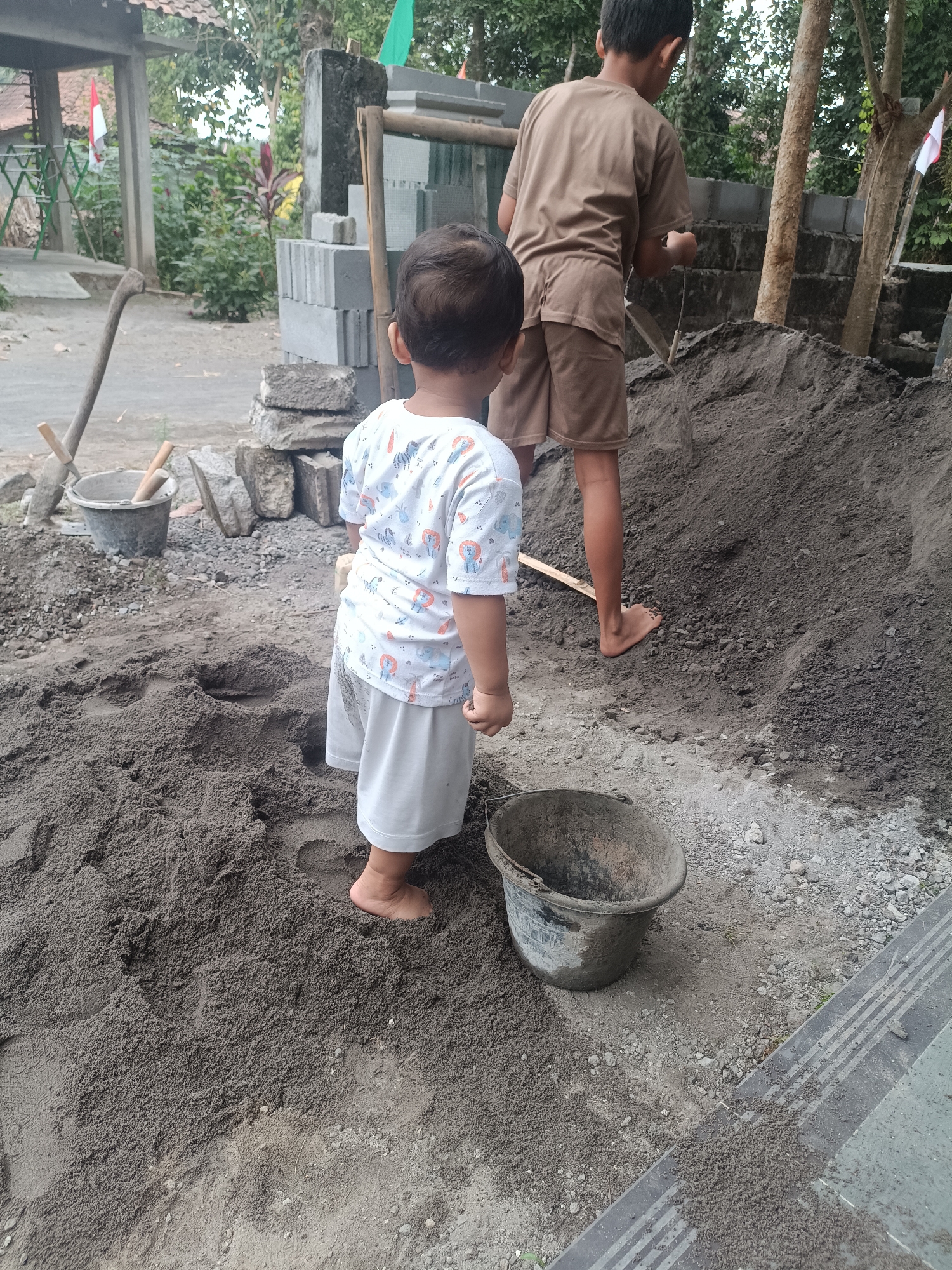 In Sumberadi, Indonesia, two young boys are engaged in a construction-related activity outdoors. The boy in the foreground, facing away from the camera, is wearing a white t-shirt with a colorful pattern and white shorts. He is barefoot and standing in a large pile of dark sand. To his right, a weathered, dark gray bucket is positioned on the ground. In the background, a slightly older boy, also facing away from the camera, is wearing a brown t-shirt and brown shorts. He is standing near another pile of sand, holding a wooden plank, and appears to be working or assisting in some capacity. The environment suggests an ongoing construction project, with stacks of gray cinder blocks visible behind the boys. Vegetation and trees are present in the background, along with a structure that may be a simple outdoor shelter. The ground is covered with sand, gravel, and dirt.