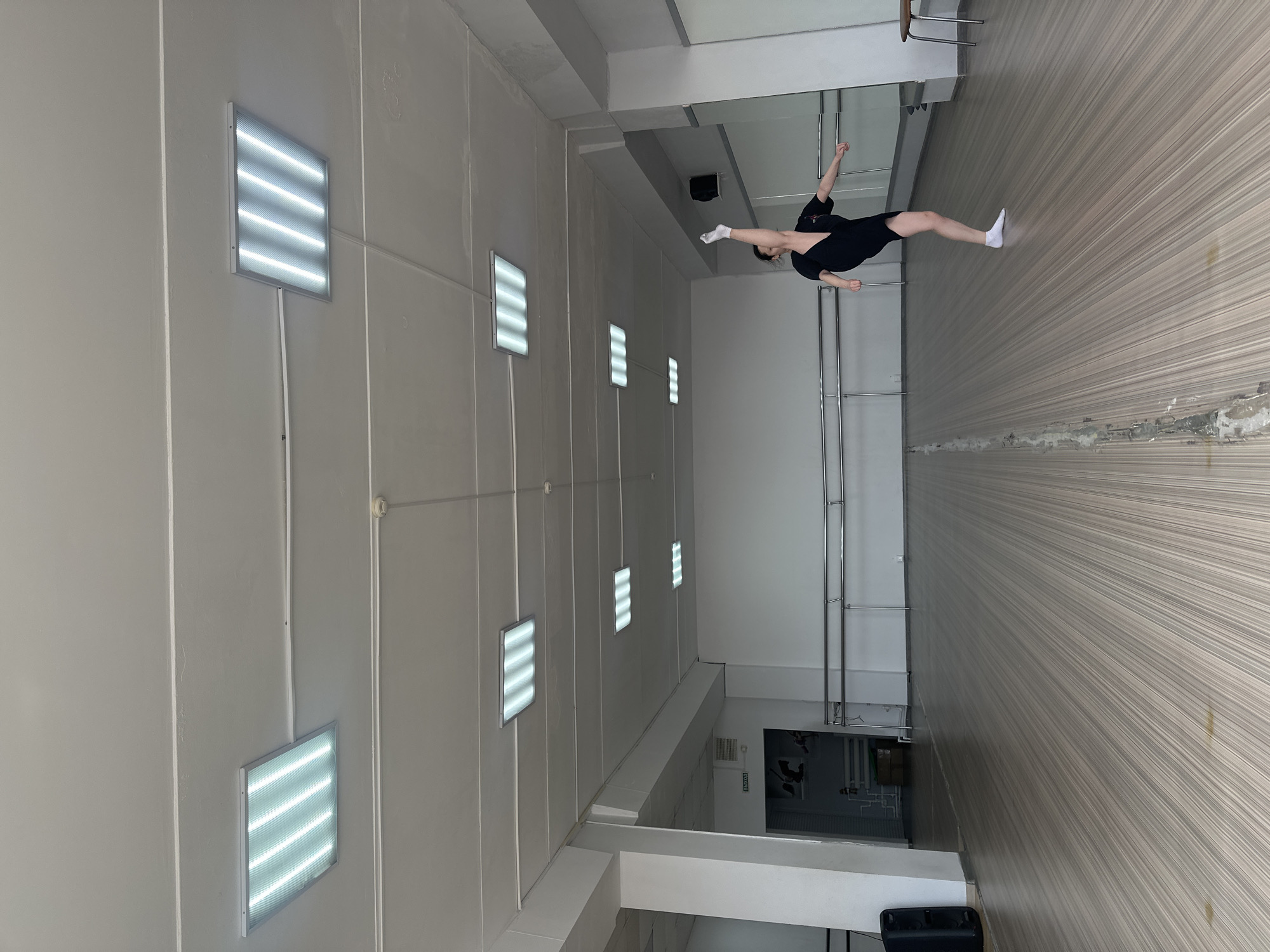 A person is in a room with a mirrored wall. The person is wearing black clothing and white socks. The location appears to be an athletic facility or dance studio with fluorescent lights on the ceiling and wood-grain flooring. There is a metal bar along the wall. The precise location in Votkinsk, Russia cannot be determined from the image alone.