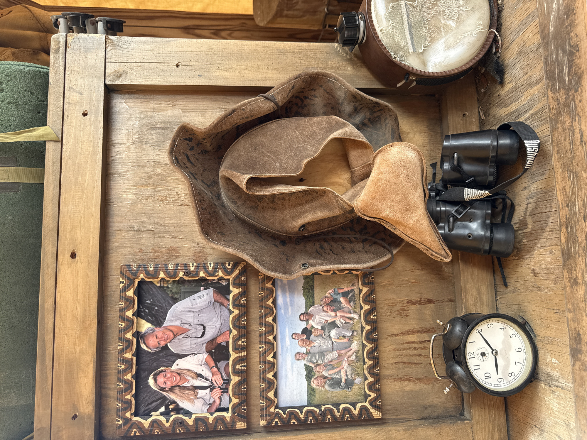 The image shows a wooden surface cluttered with various objects. Dominating the scene is a brown leather hat, partially crumpled. Beside the hat is a pair of black binoculars and a black analog alarm clock, its face displaying a time around 11:10. To the left, two framed photographs are visible. One shows a man and a woman posing, both holding cameras. The other photo features a group of approximately ten people, including adults and children, posing outdoors in a field. In the top right corner, there is a brown canteen with a light-colored cloth cover. The wooden surface itself seems to be part of a wall or shelf unit. It is possible this photo was taken in Powell, United States of America, but this cannot be confirmed with the image provided.