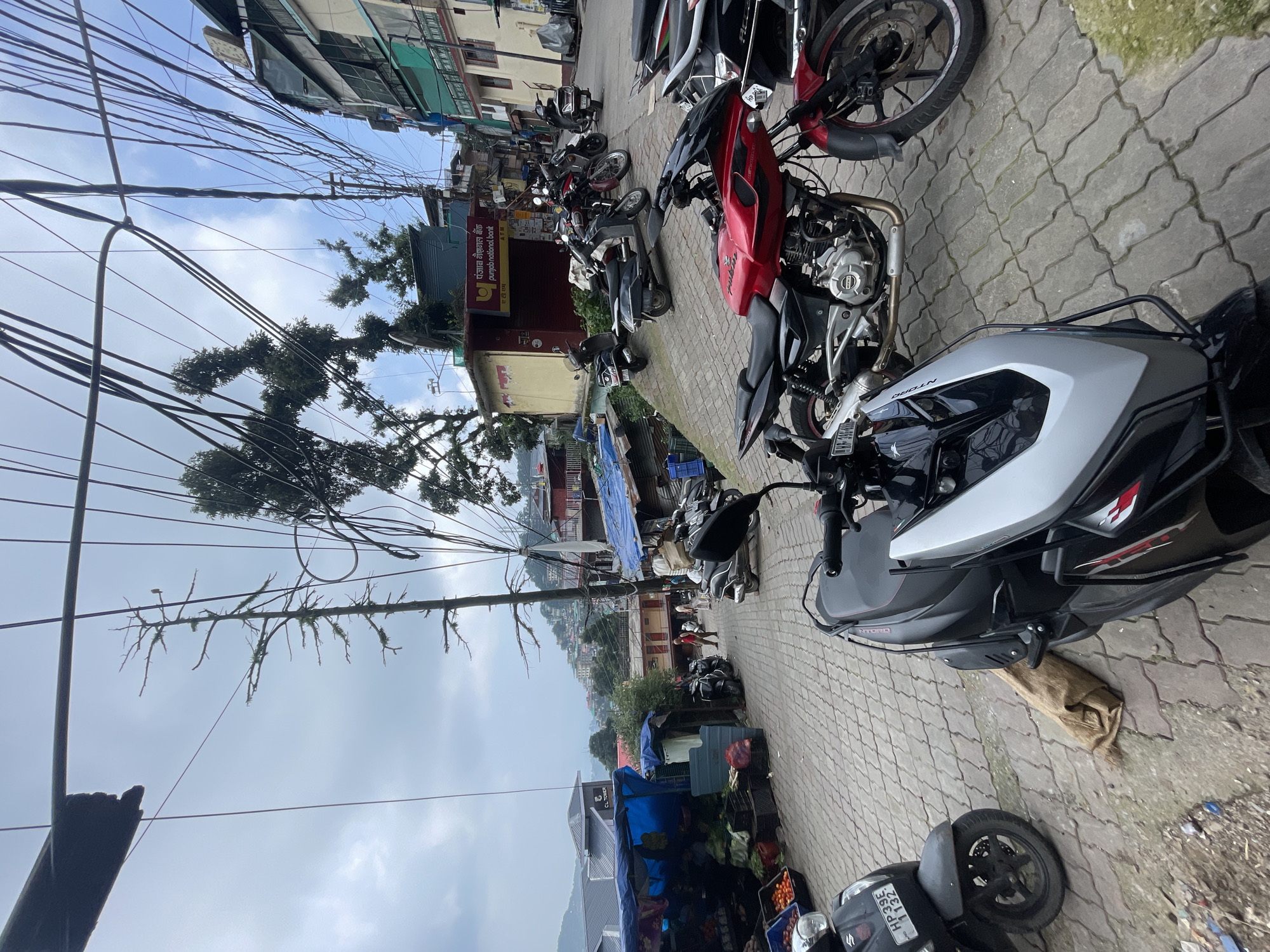 This image captures a street scene in Dharamkot, India, characterized by a narrow, winding road paved with interlocking bricks. The road is densely packed with parked motorcycles and scooters, with a few more visible further up the incline. Buildings with multiple stories and varied architectural styles line the right side of the street, adorned with numerous electrical wires crisscrossing overhead. On the left, a market stall with blue tarpaulin coverings displays fresh produce, including tomatoes and other vegetables, in crates. A few individuals are visible walking along the street. The overall impression is one of a bustling, slightly chaotic urban environment with a hilly landscape in the background, under a cloudy sky.